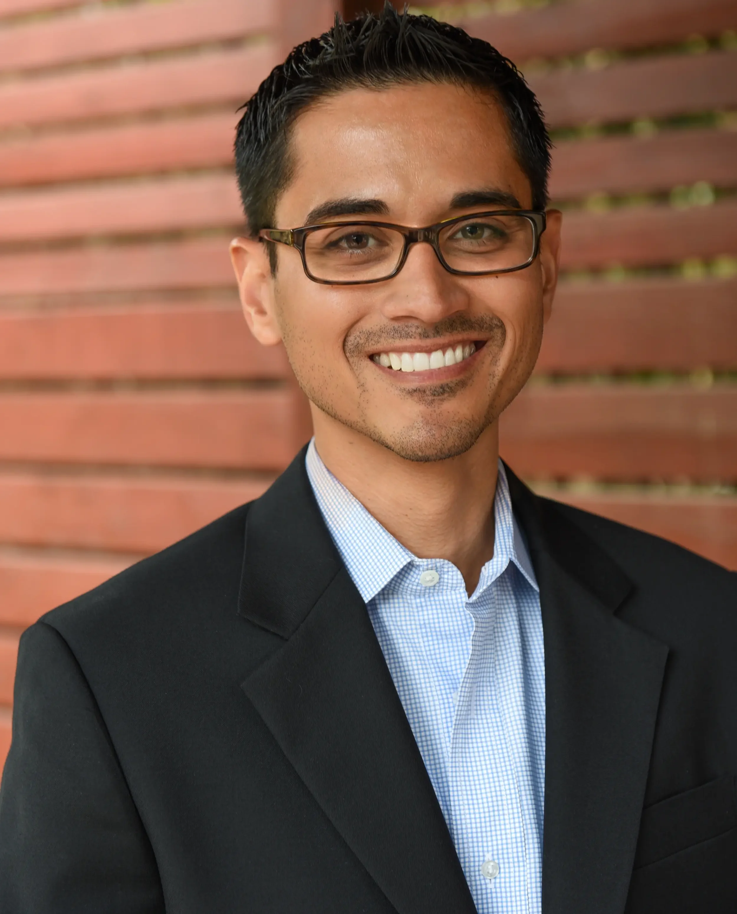Close-up of a smiling man wearing glasses, a blue checkered shirt, and a black blazer, standing in front of a background with horizontal wooden panels.