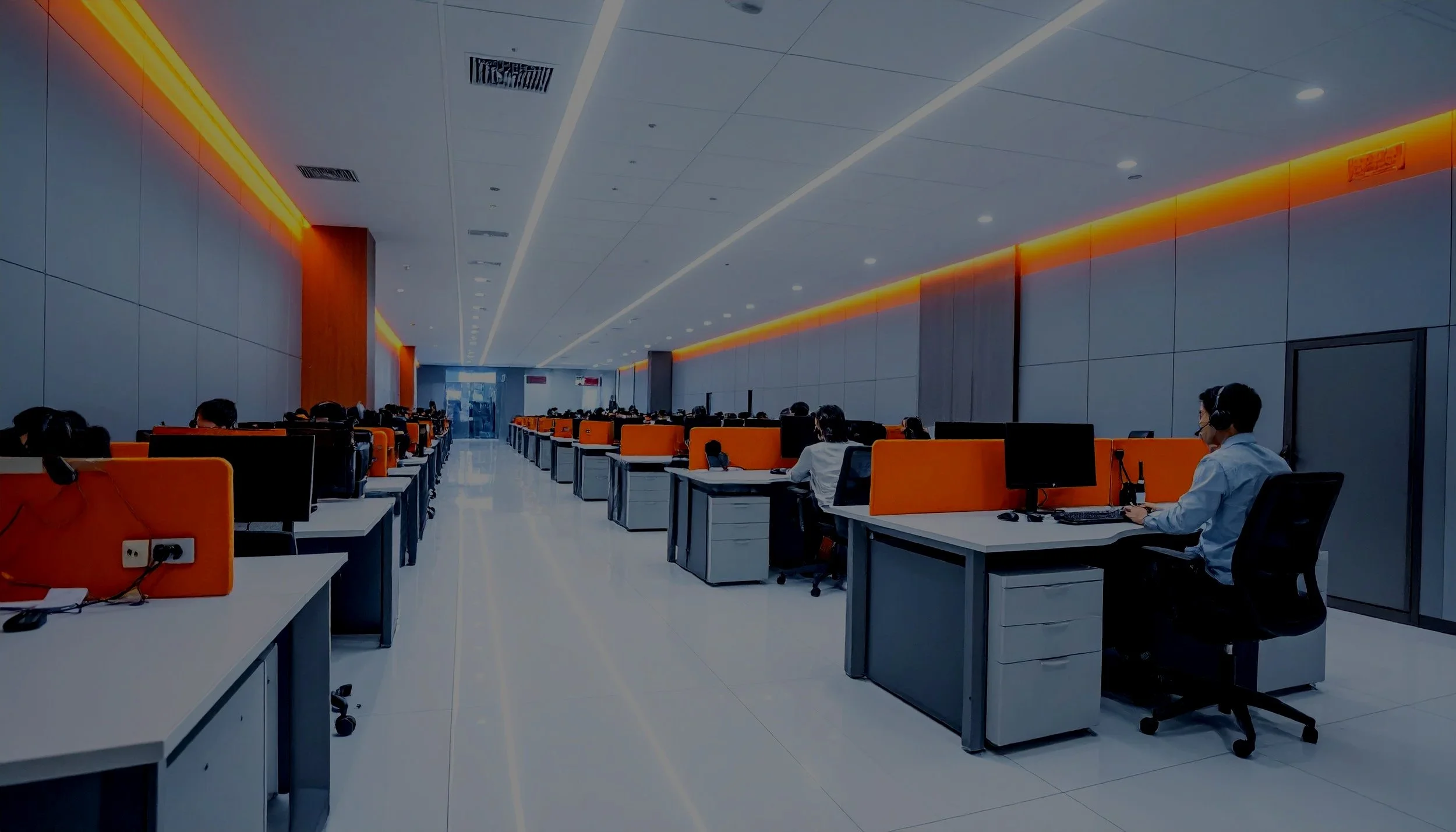 Workers sitting at desks in a modern office with orange accents and computer monitors.