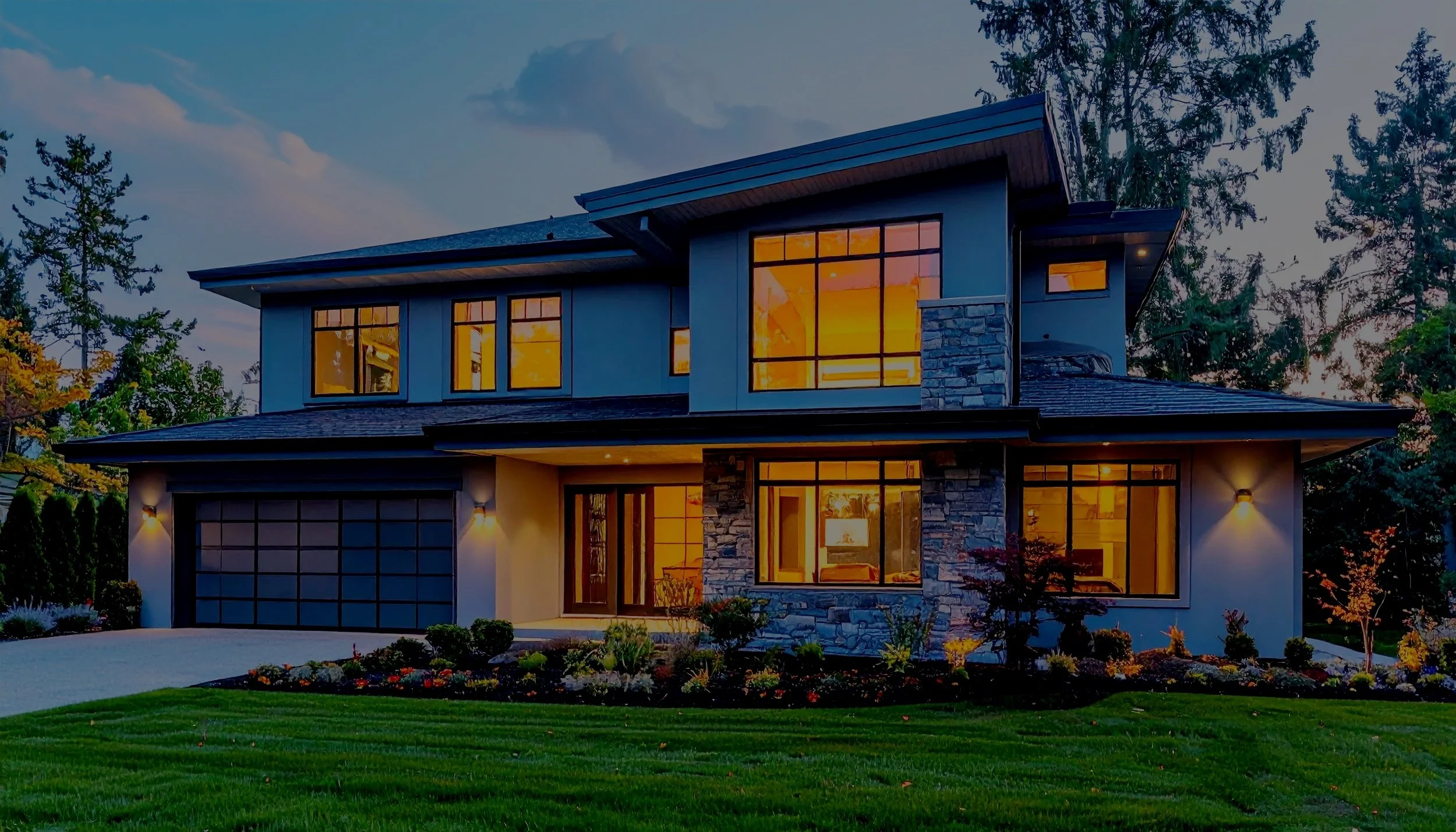 Modern two-story house with large glass windows, stone accents, and a well-maintained lawn and garden, illuminated at dusk.