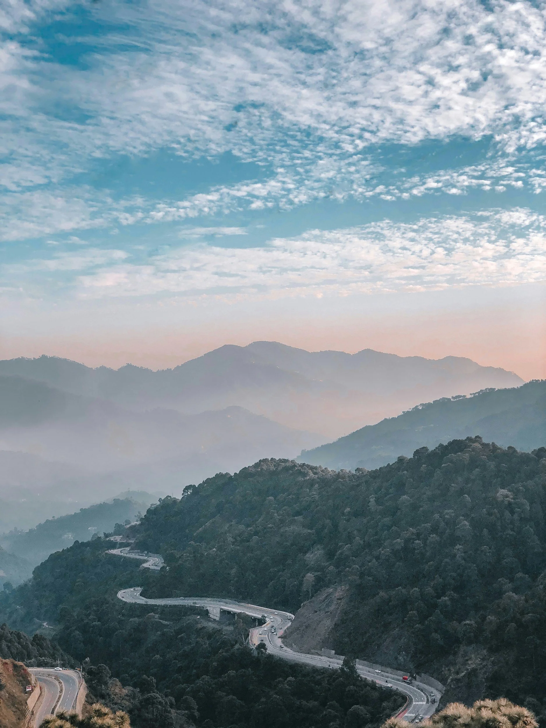 Various mountains with a winding road in a lush forest under a partly cloudy sky.