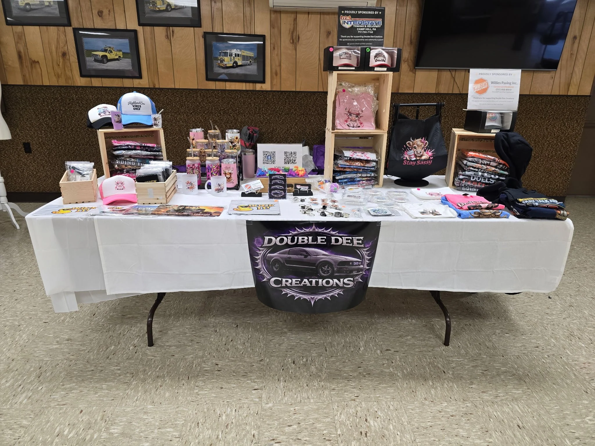 A display table with various merchandise including tumblers, hats, mugs, T-shirts, buttons, and other items, set up at an indoor event or market space. Behind the table, there are framed pictures of vehicles on a wooden wall.