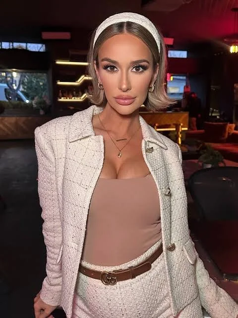 A young woman with blonde hair, wearing a white headband, beige top, matching white blazer and pants, and a brown belt, posing indoors with a sophisticated background.