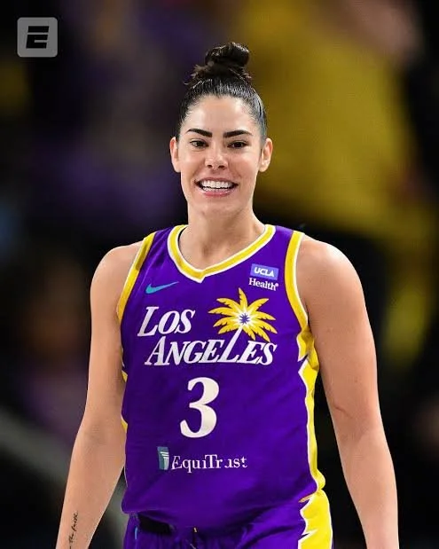 A female basketball player wearing a purple Los Angeles basketball jersey with the number 3, smiling, with her hair tied up in a bun during a game.