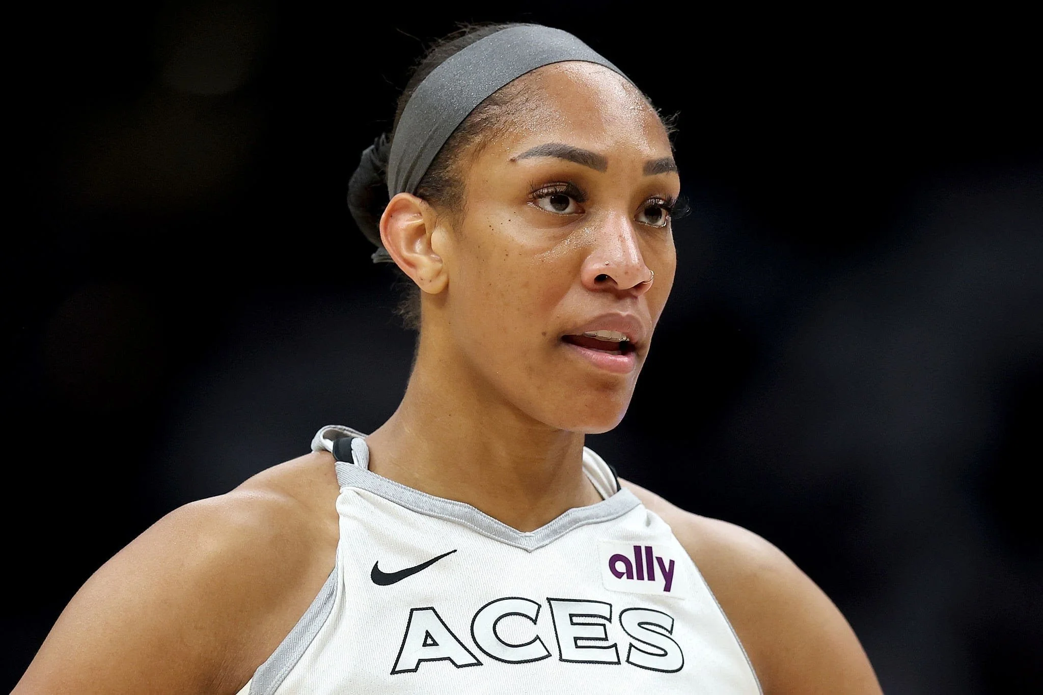 A female athlete with a focused expression wearing a white sports jersey with the words 'ACES' and sponsor logos, a gray headband, and sweat visible on her face.