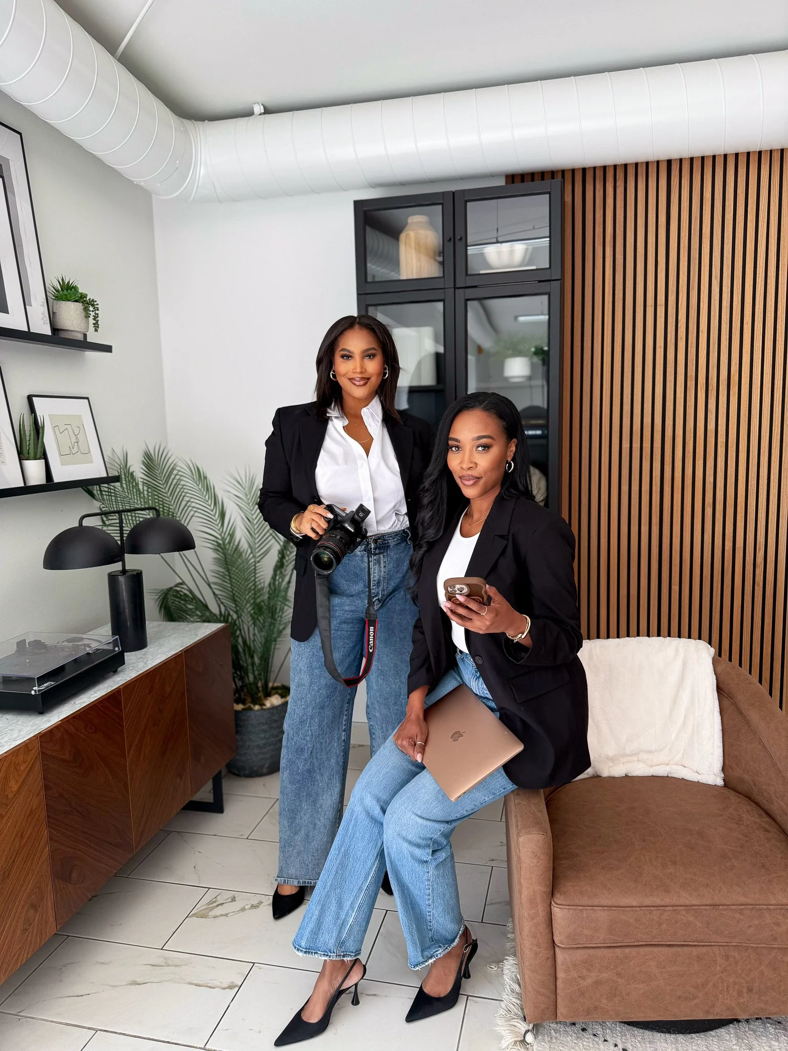 Two women in business attire in an office, one sitting with a laptop and phone, the other standing with a camera, office decor includes a wooden wall, indoor plants, and framed pictures.