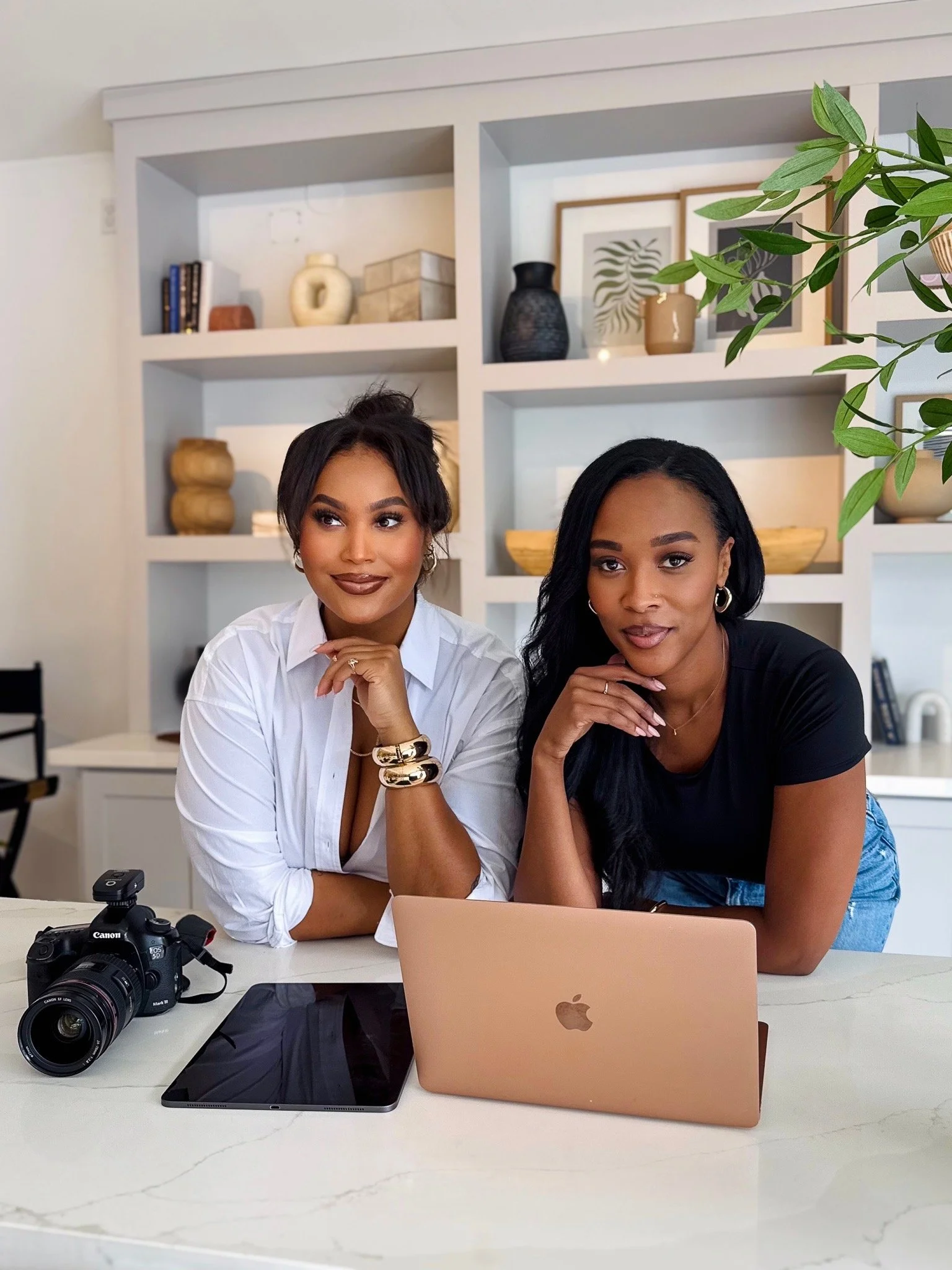 Two women sitting at a table with a laptop, camera, and tablet in a modern living room.