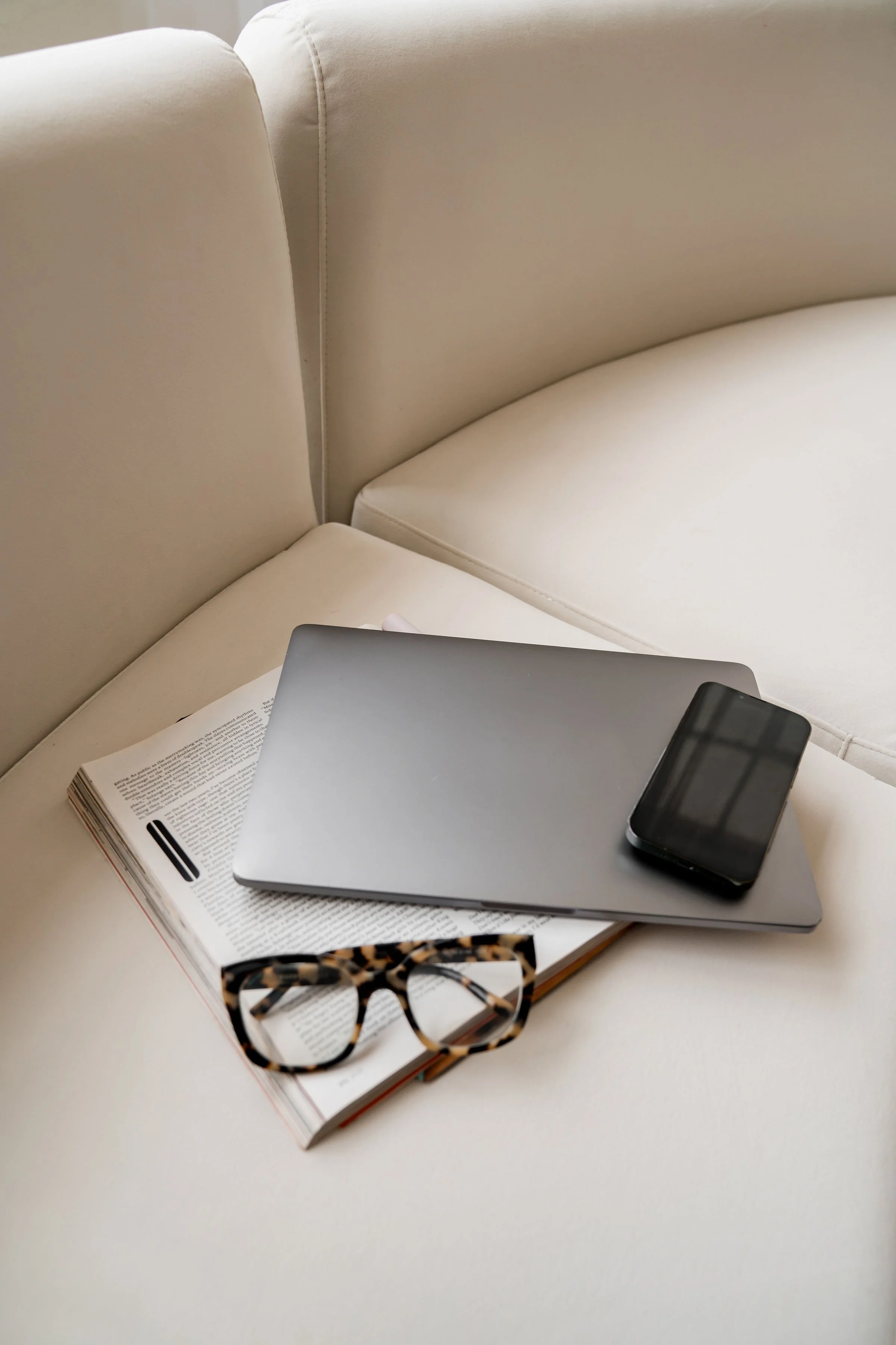 A laptop, smartphone, reader, and reading glasses arranged on an open book on a white sofa.