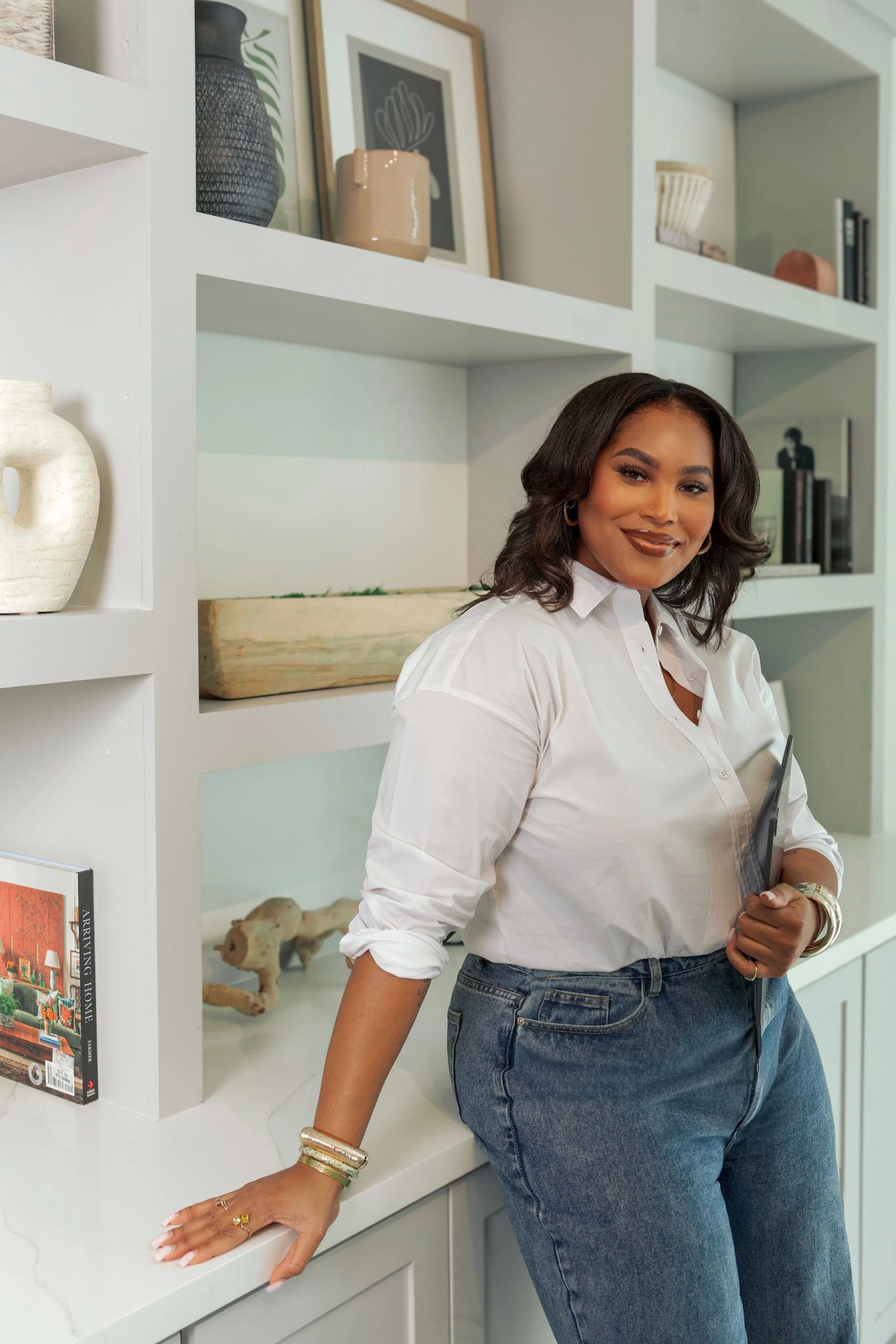 A woman with dark hair, wearing a white shirt and blue jeans, standing in front of a white bookshelf with decorative items and books, smiling at the camera.