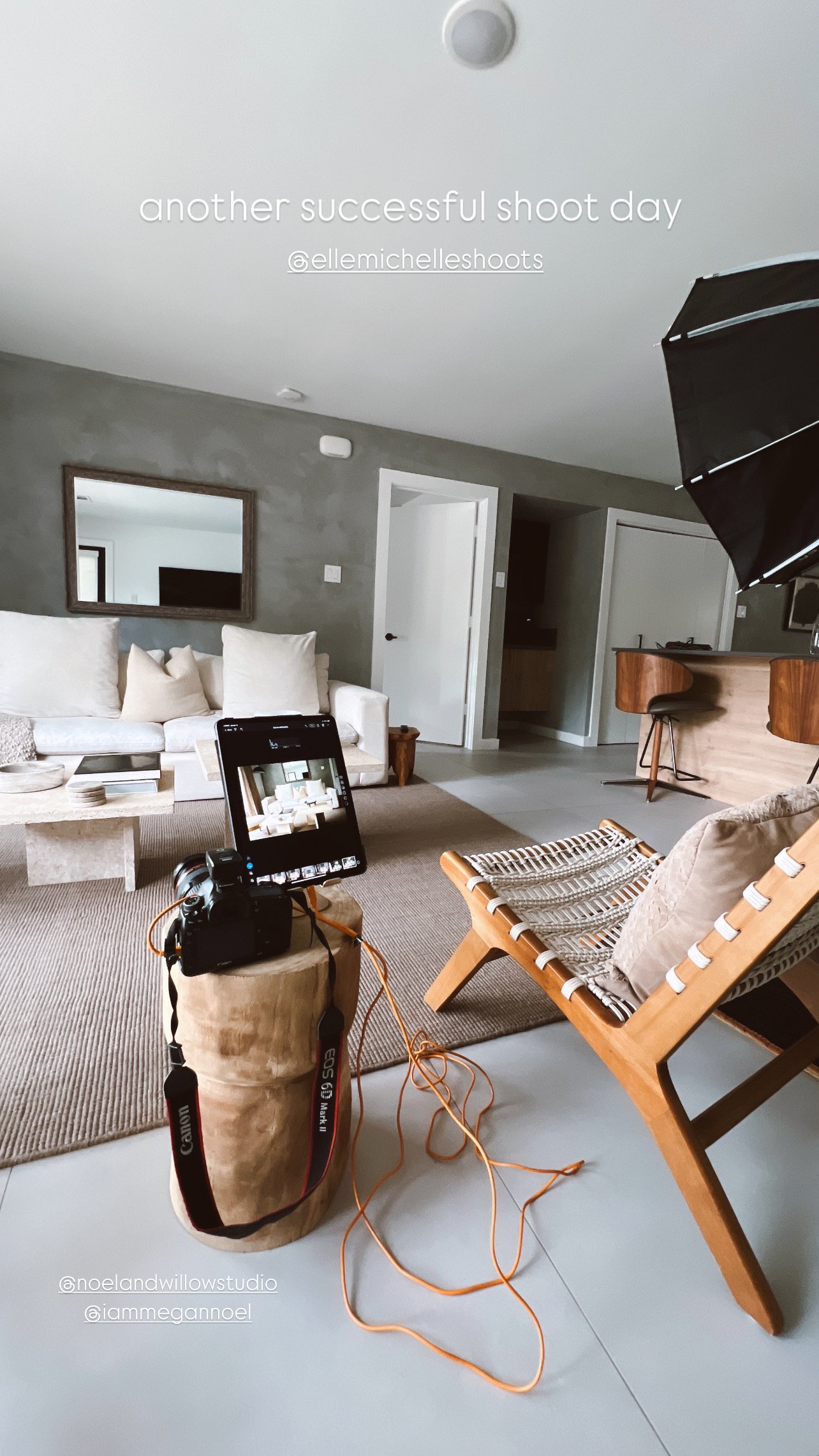 Living room with camera and monitor set up for a photo shoot, decorated in neutral tones, with seating and bar area in background.