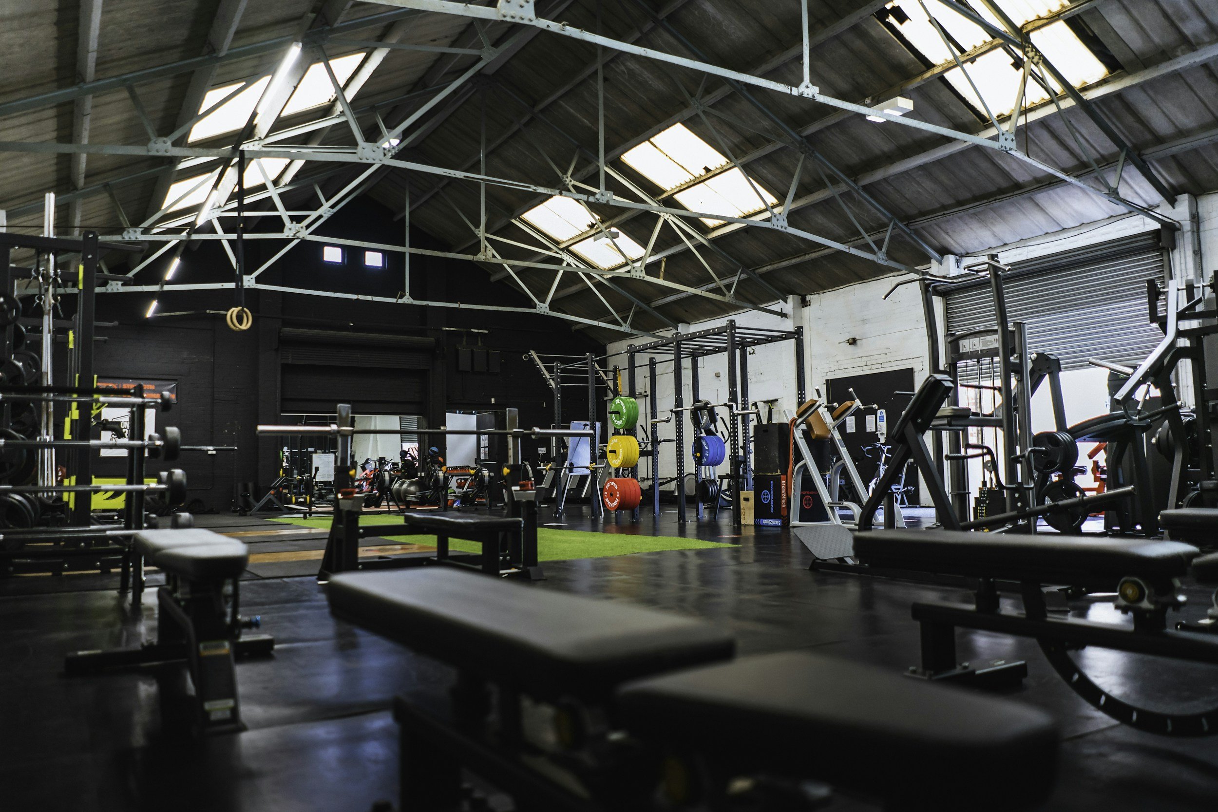 An empty gym with various workout equipment, including benches, weight racks, and colorful weights, inside a spacious industrial-style building with a high ceiling and skylights.