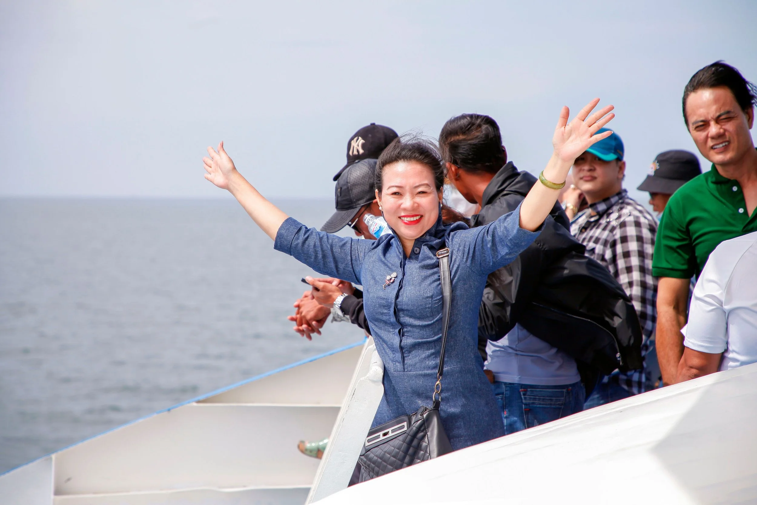 A smiling traveler raises her arms in excitement aboard a boat, with fellow passengers and the sea in the background.