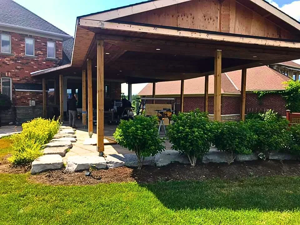 Backyard with a covered wooden patio, stone pathway, green bushes, and a grassy lawn in front of a brick house.