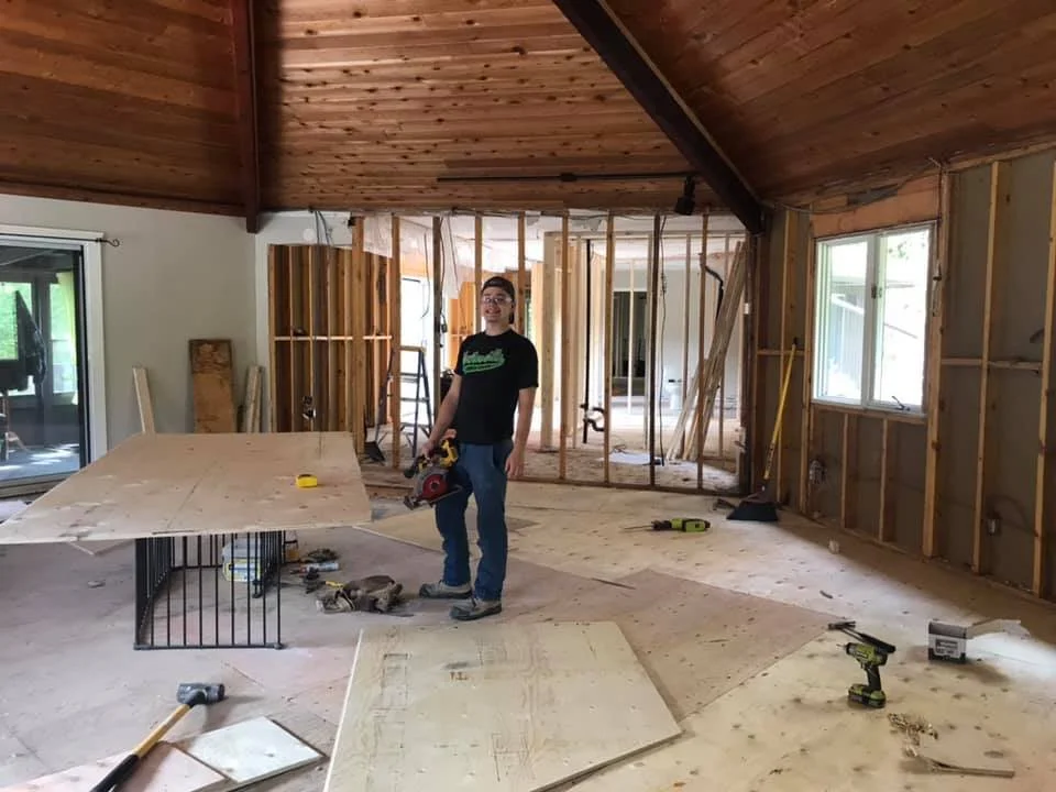 Taylor standing inside a home under construction or renovation, holding a power saw, with unfinished framing and construction tools scattered around.