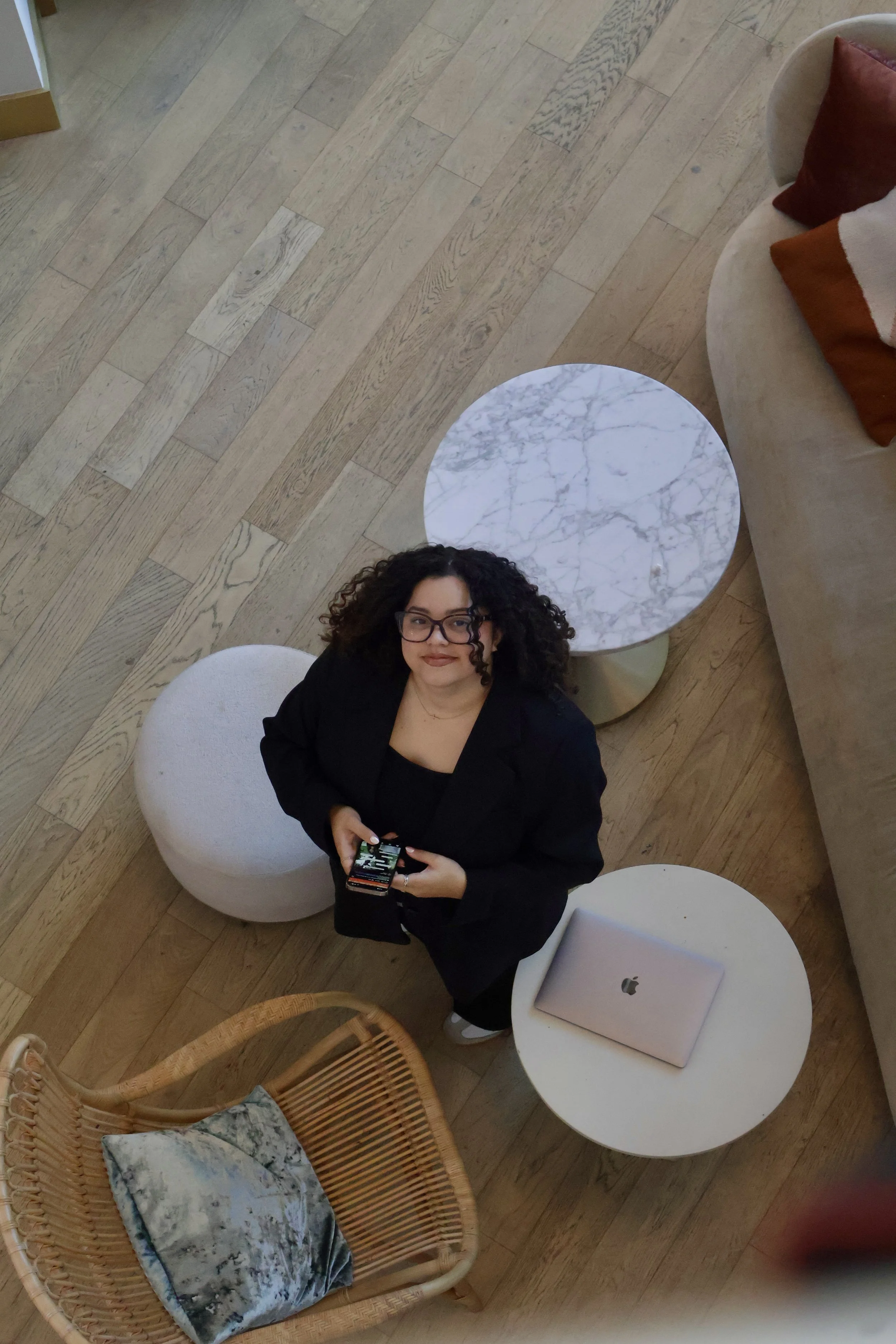 A woman with curly hair and glasses standing in a modern living room, holding a smartphone, surrounded by a round marble coffee table, a white ottoman, a wicker chair with a cushion, and an Apple MacBook on a small round table, with hardwood flooring and a beige couch with red and brown pillows nearby.