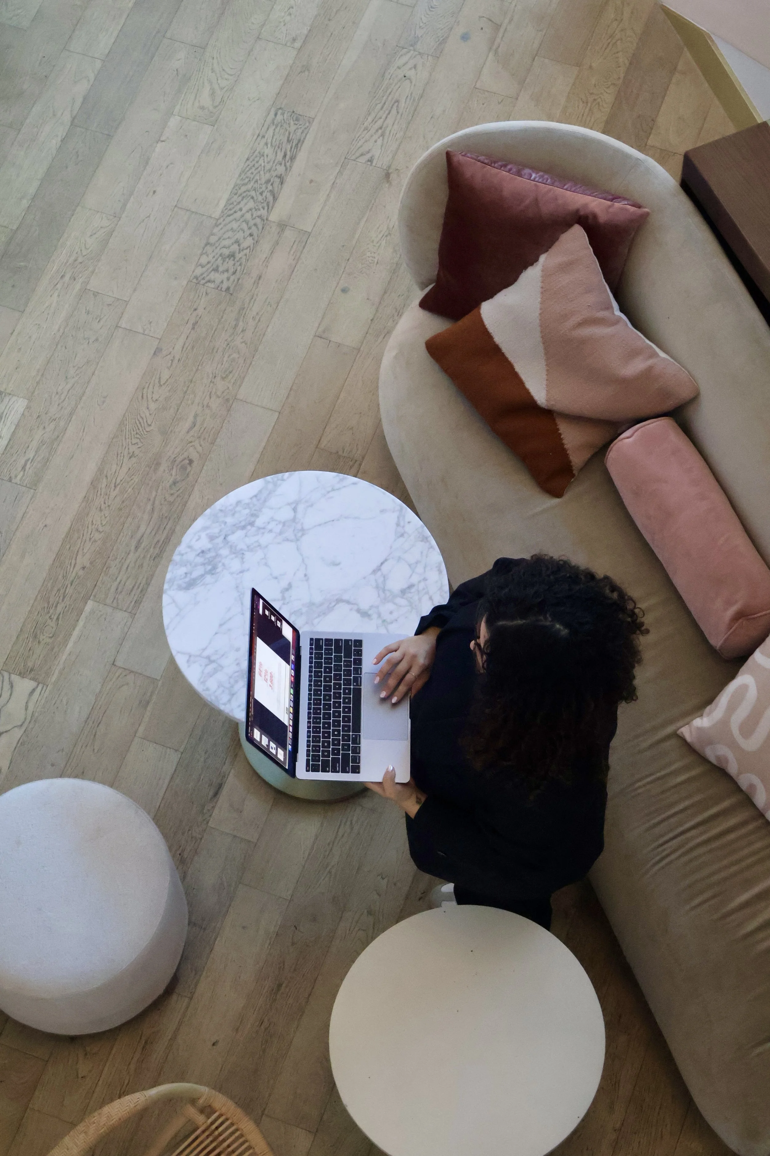 A woman with curly hair using a laptop at a small round marble table in a cozy living room with a beige sofa and pastel-colored pillows, hardwood floor, and minimalist decor.