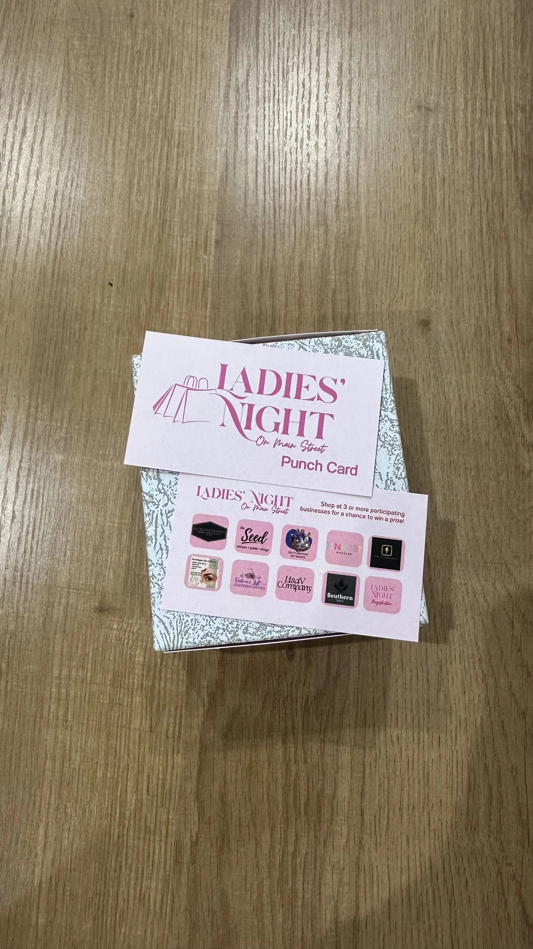 A pink ticket and a pink card on top of a silver gift box on a wooden table, promoting a Ladies' Night event with participating businesses and a chance to win a prize.
