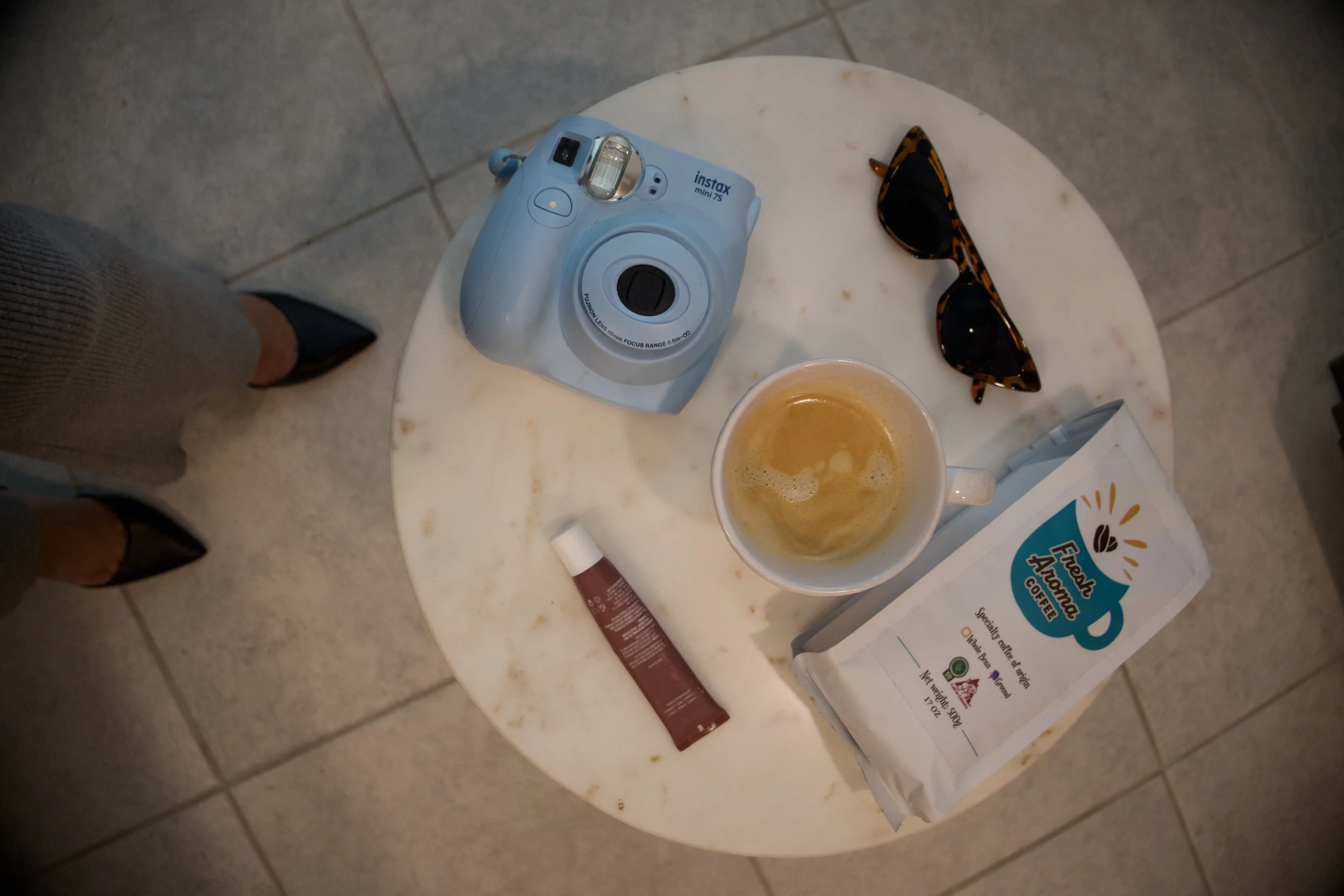 Overhead view of a round marble table with a blue instant camera, a pair of tortoiseshell sunglasses, a mug of coffee with foam, a standing bag of coffee, a small tube of cream, and a person's black high heel shoes and gray pants.