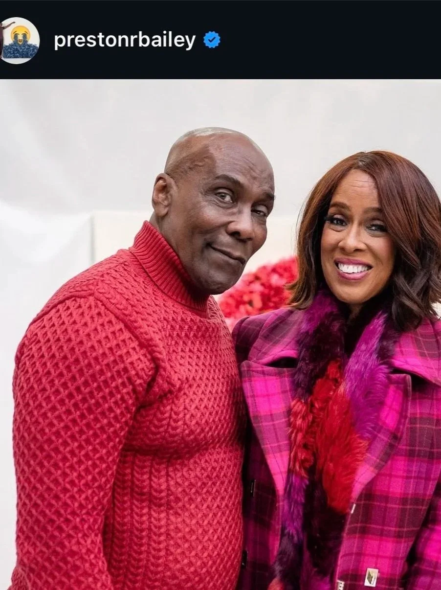 Two people, a man and a woman, standing close together, smiling, with a blurred background and red flowers visible.