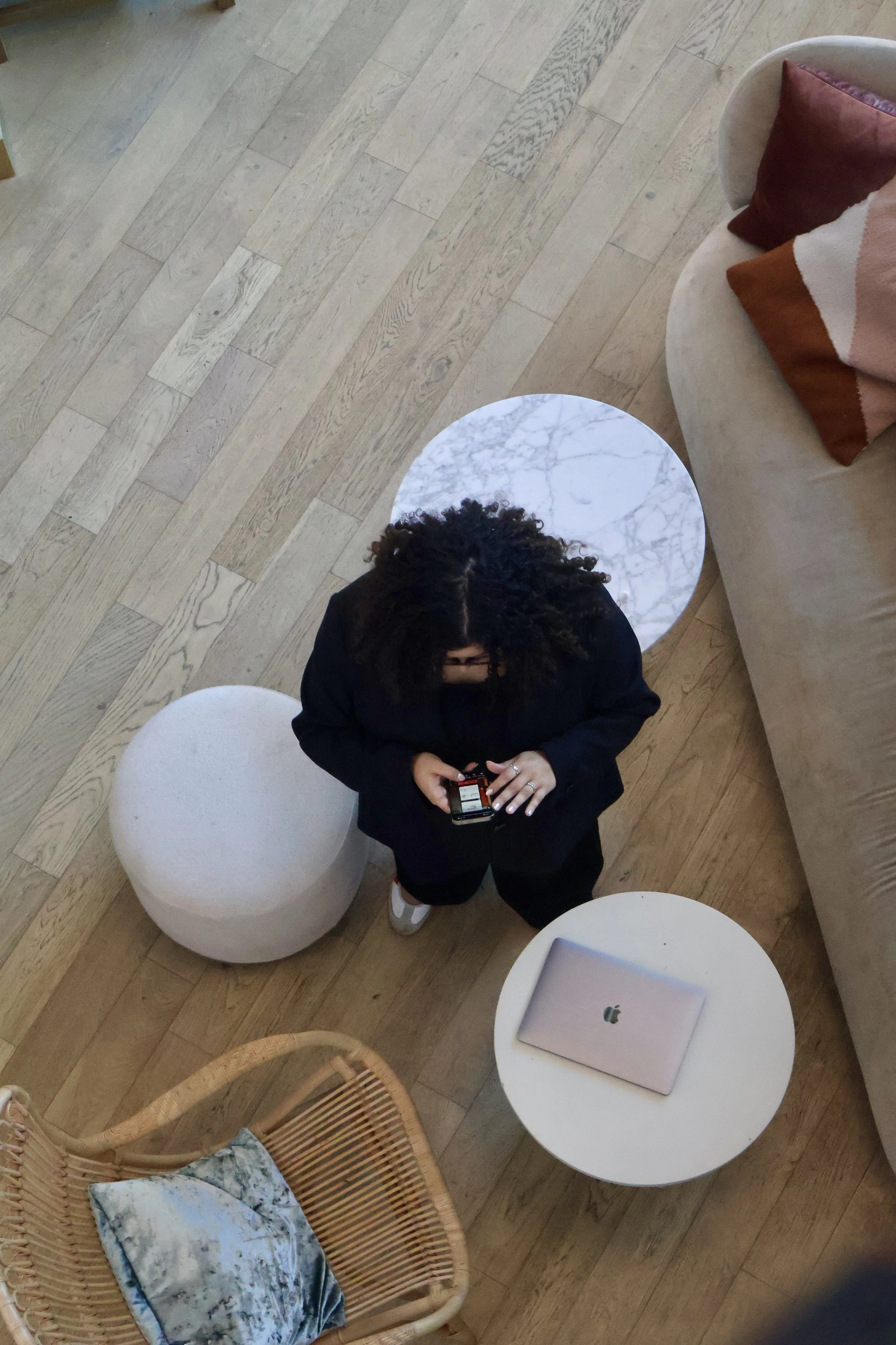 A person with curly hair looking at a smartphone in a living room. There is a small white round table with a laptop, a round white ottoman, a beige sofa with cushions, a rattan chair with a patterned fabric, and a marble-topped table on a wooden floor.