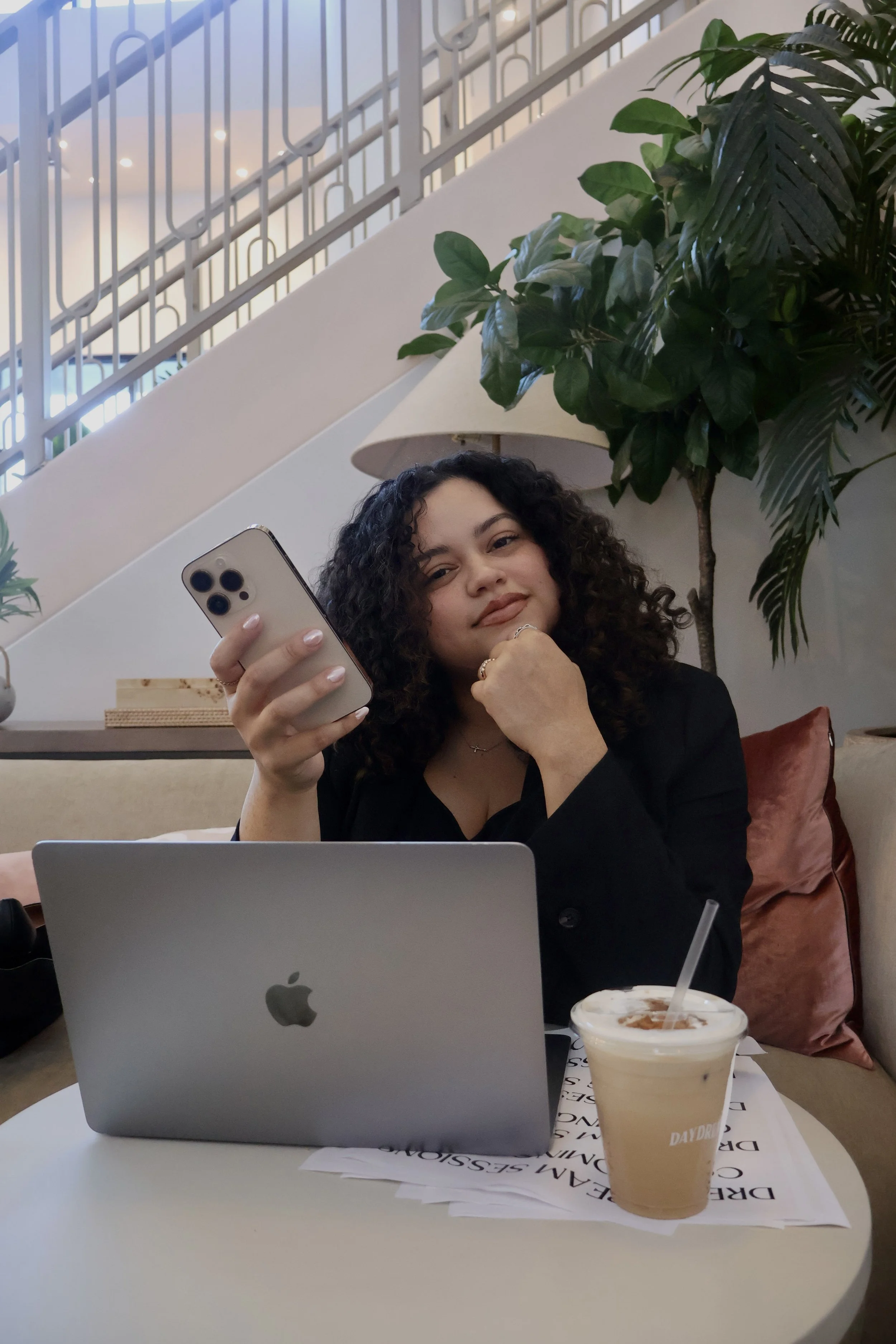 A young woman with curly hair sitting at a table in a cafe, holding a smartphone in her right hand, with a laptop open in front of her. There is a cold coffee drink on the table, along with some papers. Behind her, there are large green plants and a staircase with a metal railing.