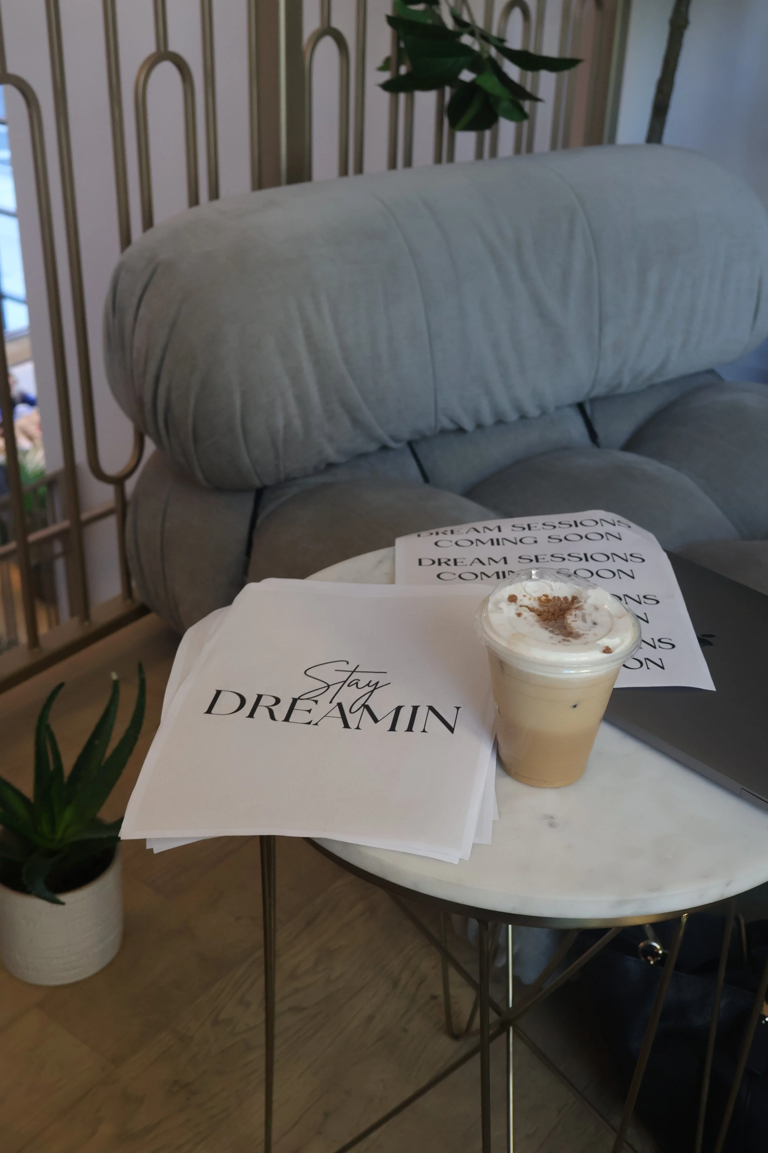 A small round marble-topped table in a cozy cafe. On the table are printed papers with the quote 'Stay Dreamin,' a cup of iced coffee with whipped cream and cocoa powder, a closed laptop, and a potted succulent plant nearby. A gray upholstered sofa and a decorative metal railing are in the background.