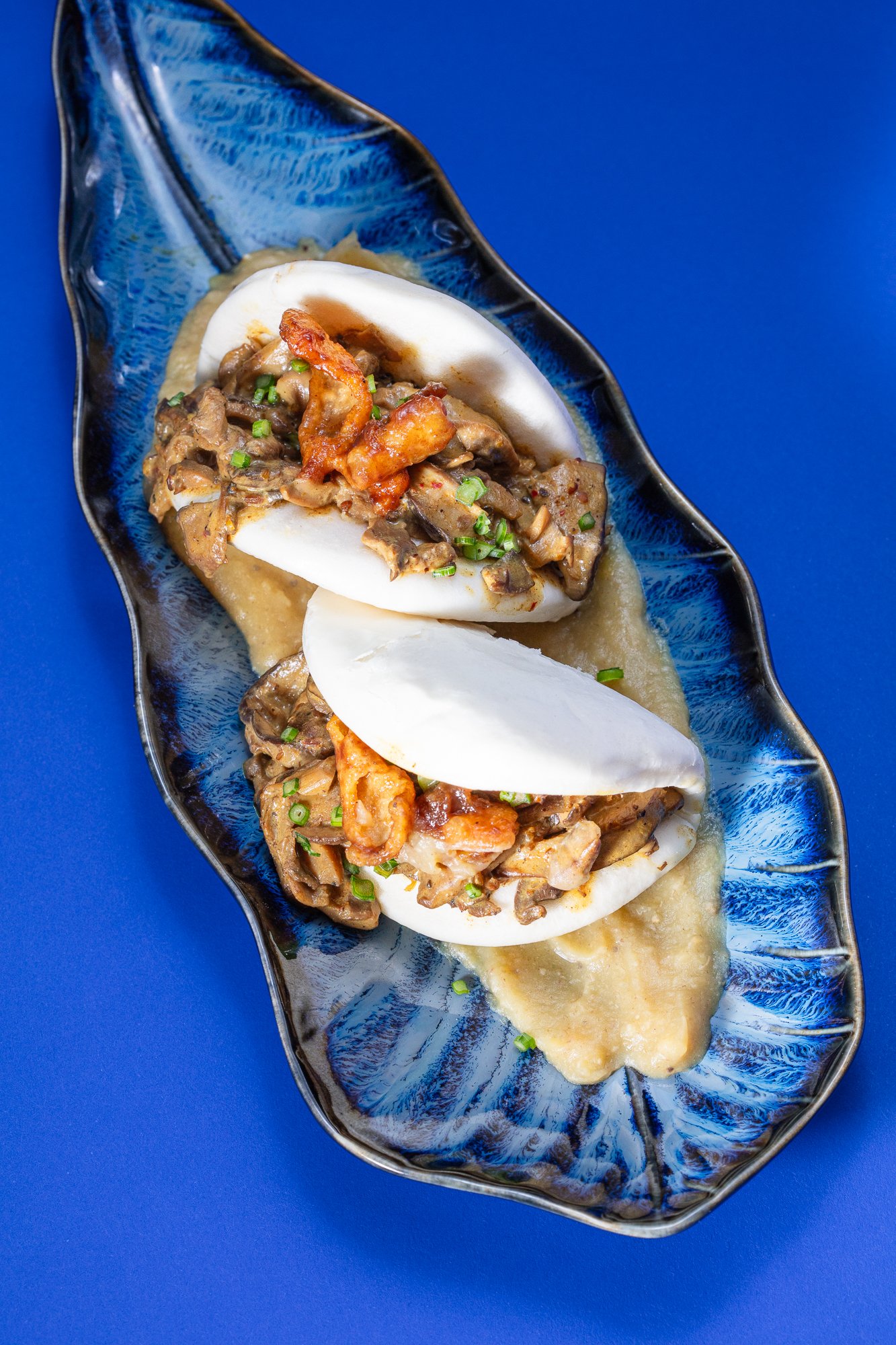 Two steamed bao buns filled with stir-fried mushrooms and chicken, topped with chopped green onions, served on a black leaf-shaped platter with sauce underneath, against a blue background.