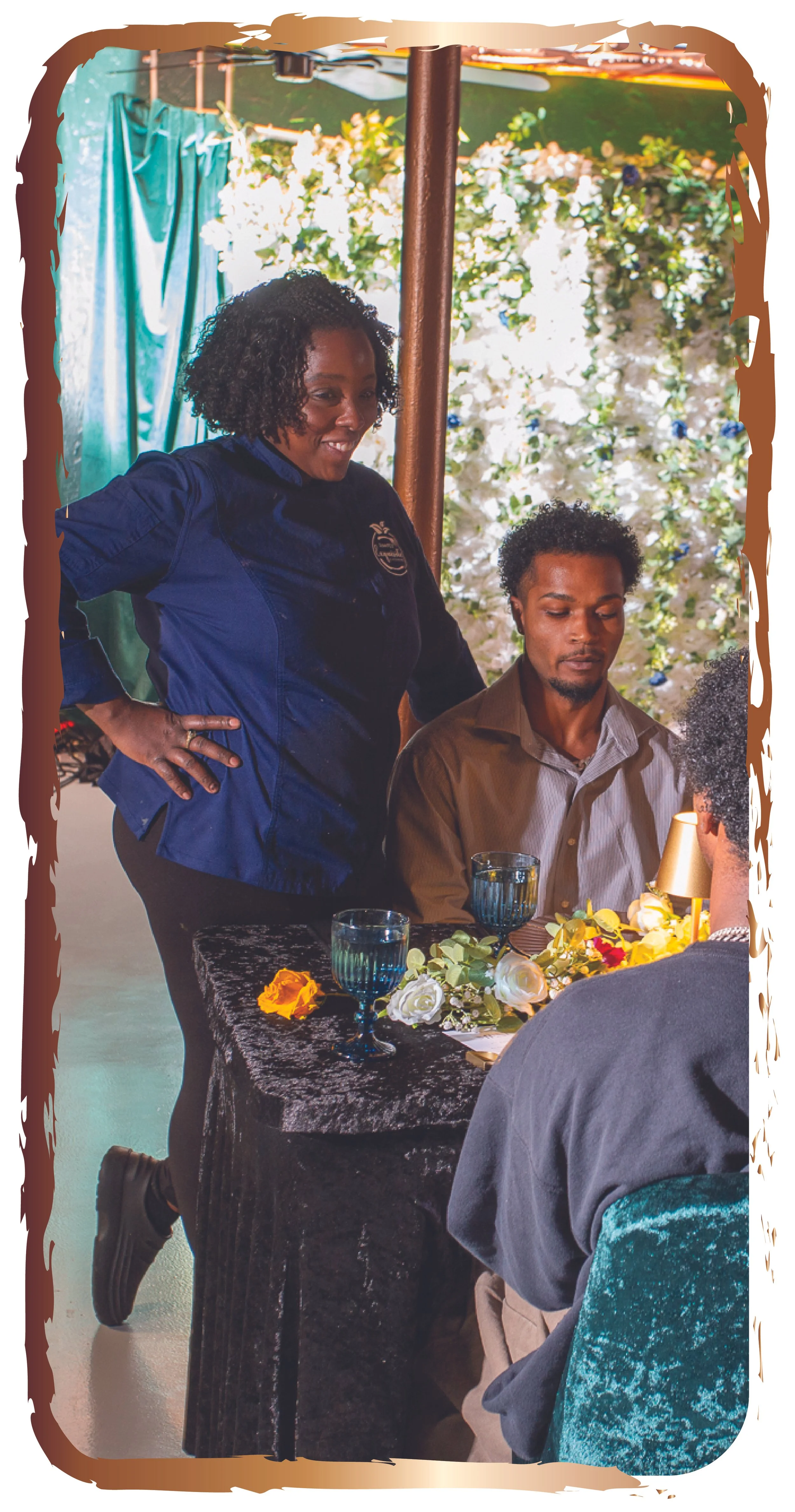 A woman in a navy chef coat standing next to a seated man at a table decorated with flowers and candles, in a setting with floral background and teal curtains.