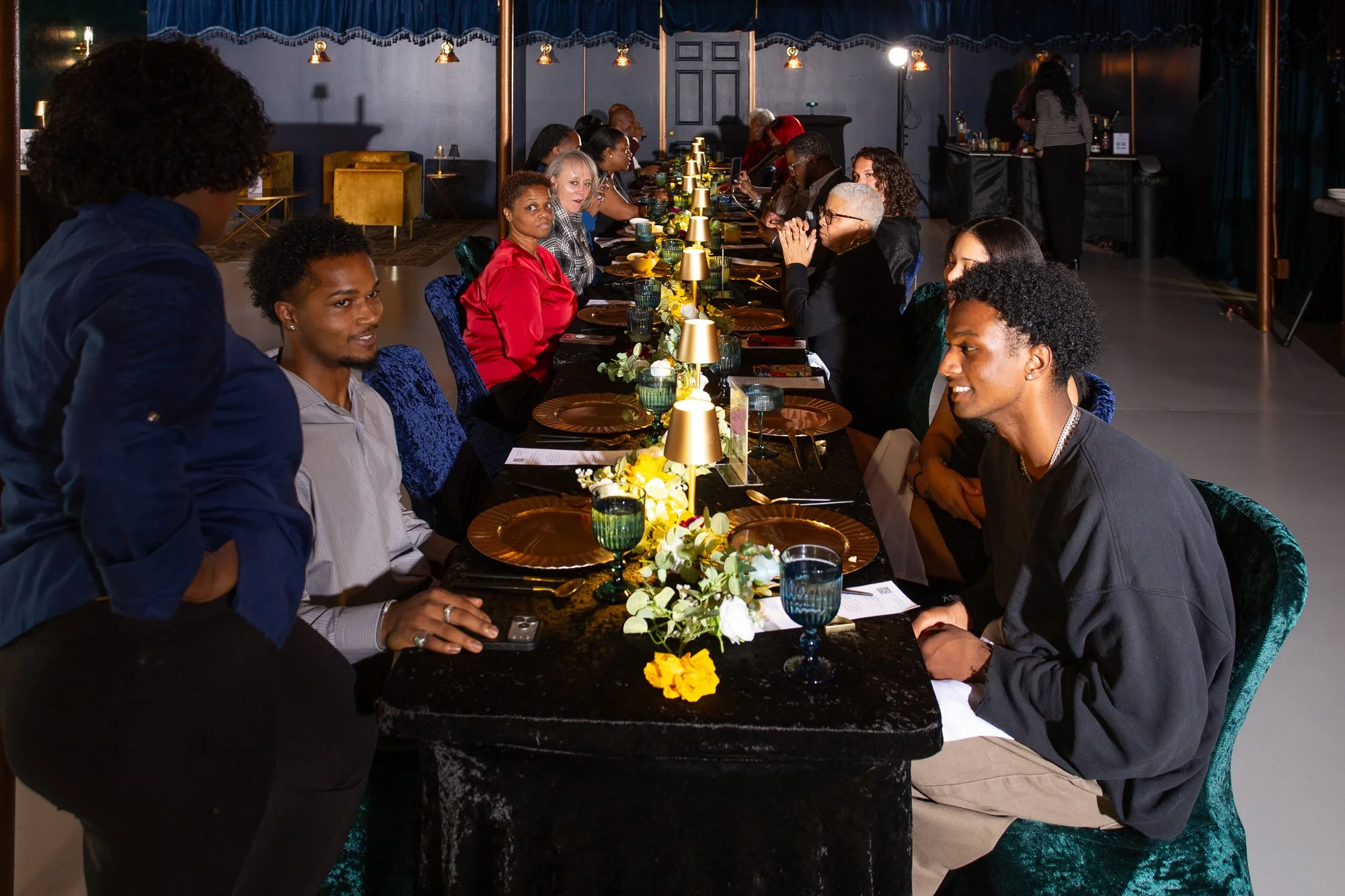 A group of people sitting at a long dining table with elegant tableware and floral decorations, engaging in conversation at a formal event or dinner gathering.