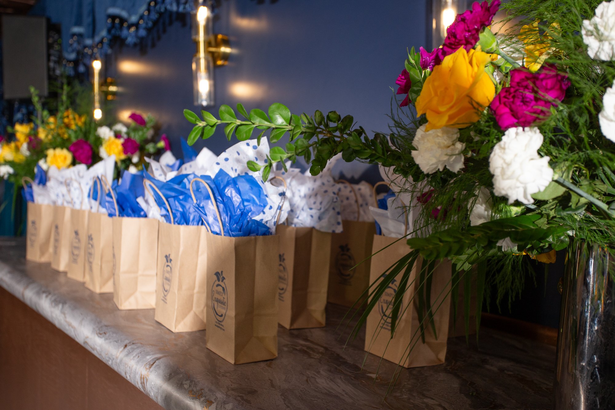 Arranged gift bags with blue and white tissue paper and a large bouquet of colorful flowers including yellow, pink, white, and greenery, on a marble countertop.