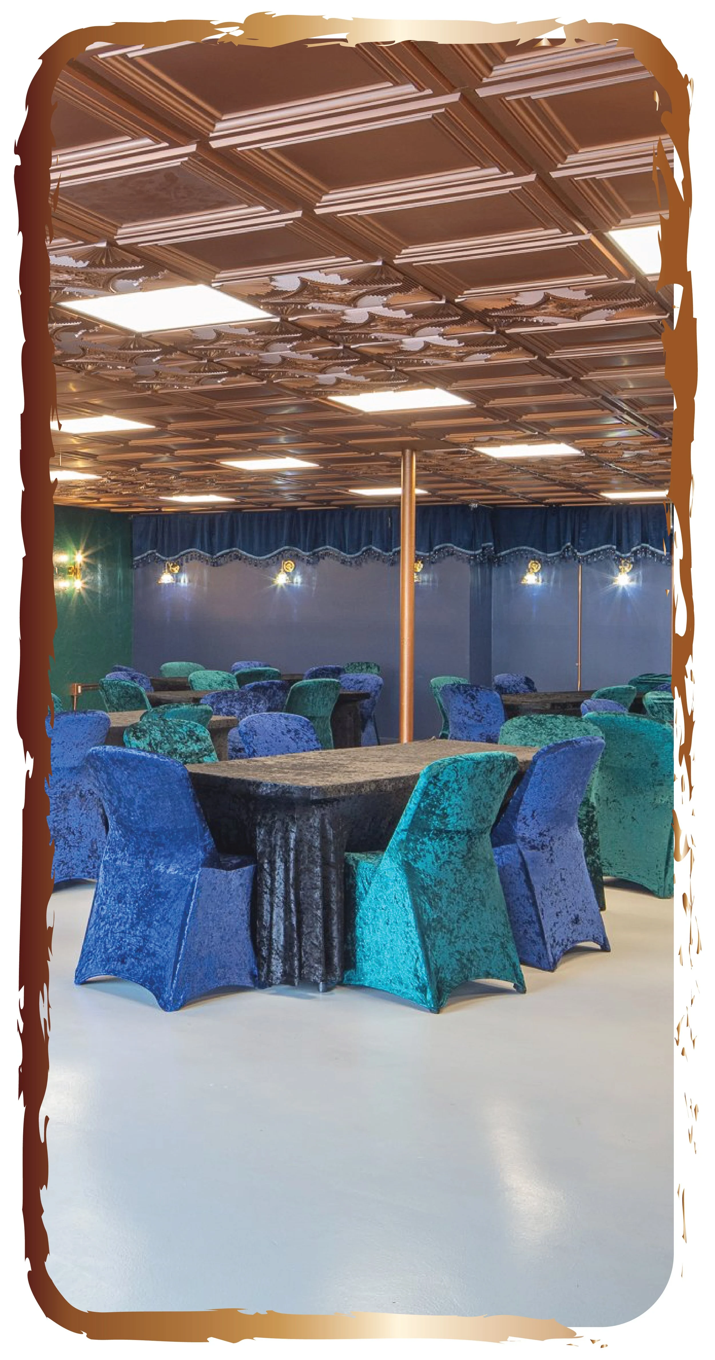 An elegant event space featuring multiple round tables covered with black velvet tablecloths. The chairs are draped in matching velvet covers in shades of blue and teal. The ceiling has a decorative wooden pattern with lighting fixtures, and there are blue curtains along the back wall with wall-mounted lights, creating a sophisticated ambiance.