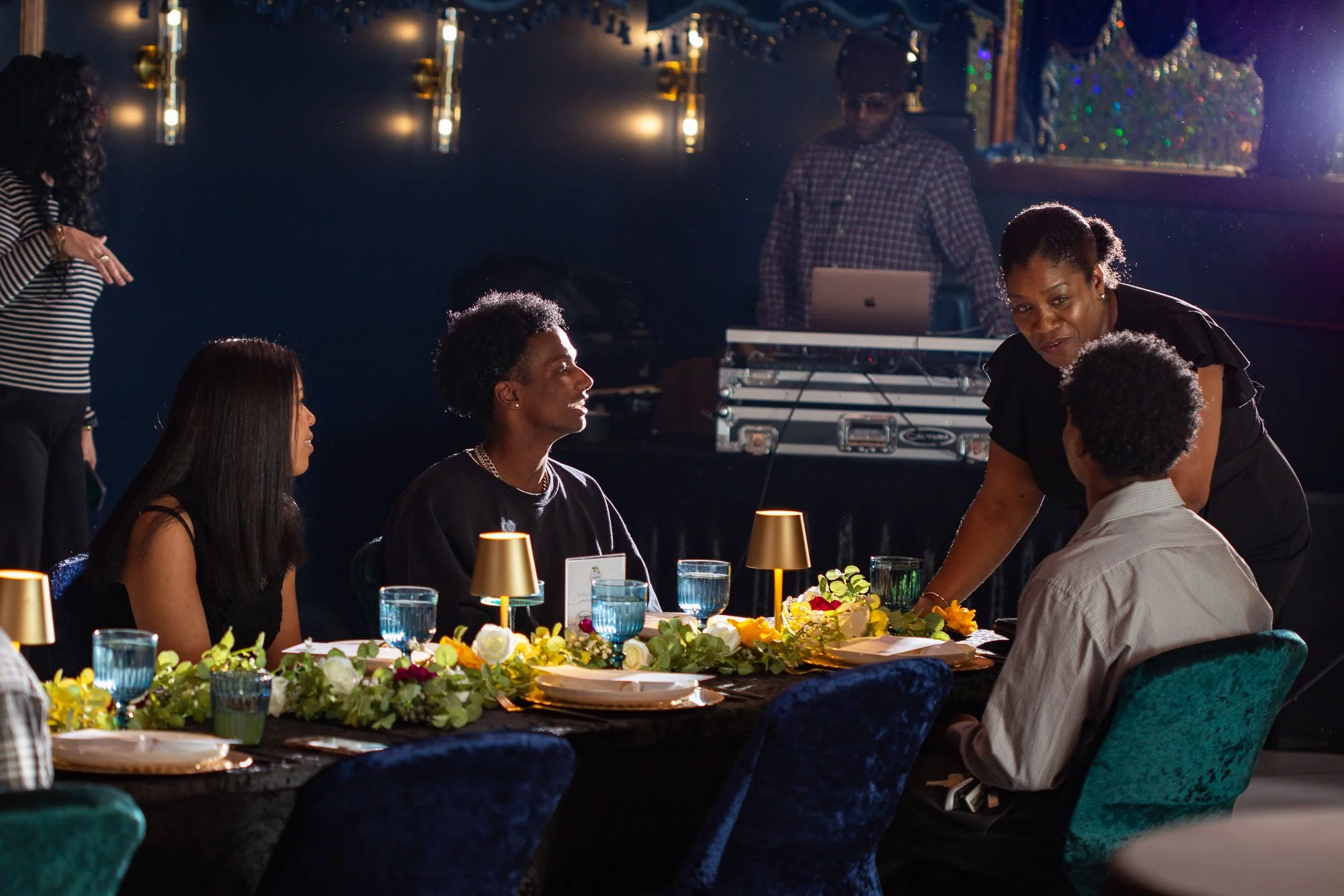 A group of people sitting around a festively decorated table at an upscale event, with a woman standing and speaking to them, and a DJ in the background.