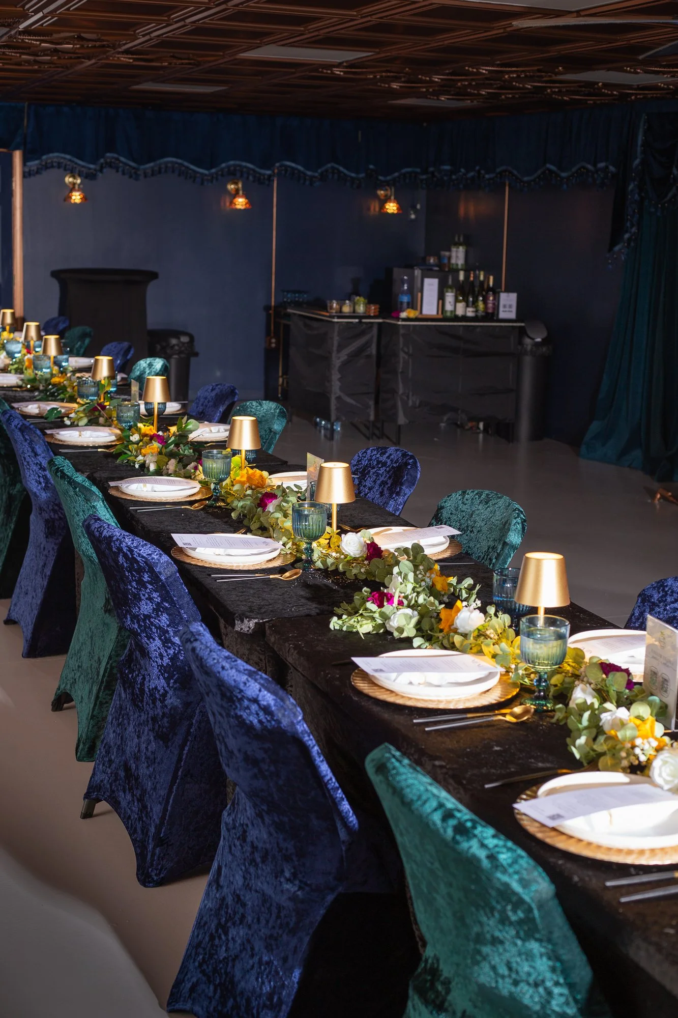 A long banquet table set for a formal event with colorful velvet chairs, gold lamps, floral centerpieces, and place settings with plates, napkins, and cutlery. The background features a dark blue curtain and a bar area.