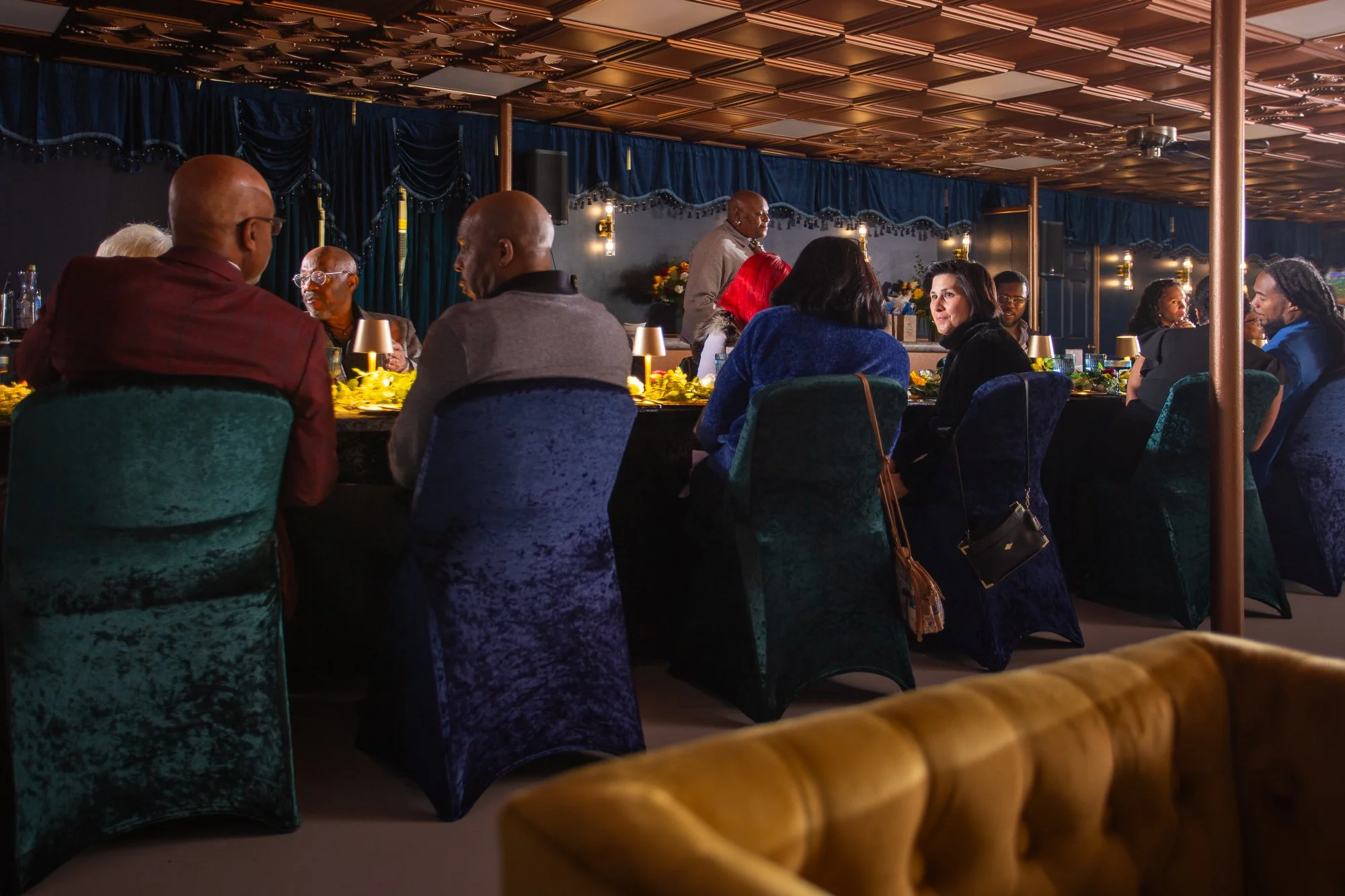 People seated around a dinner table at an indoor event, with some engaging in conversation and others looking around, in a dimly lit decorated venue with blue velvet curtains and lamps.