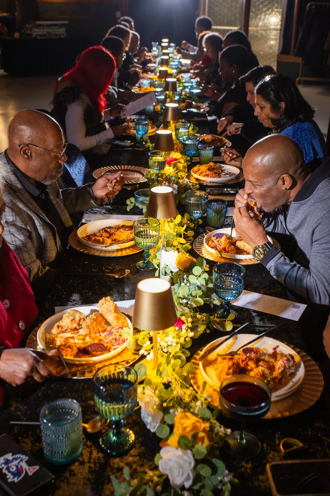 People gathered around a long dining table eating a meal during a dinner event. The table is decorated with floral arrangements, candles, and blue glassware.