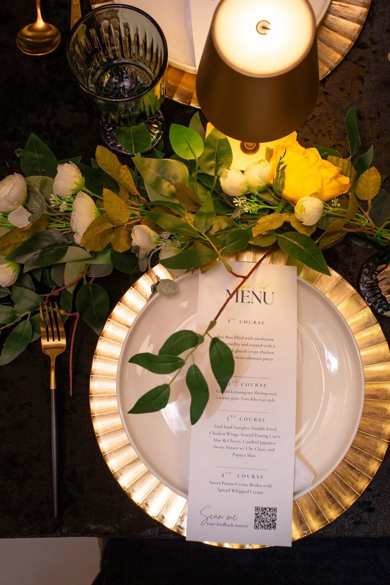Elegant table setting with a white plate on a gold-rimmed charger, decorated with a floral arrangement of white and yellow flowers, green leaves, and a menu card. A gold fork is placed to the left of the plate, and there is a glass and a candle on th