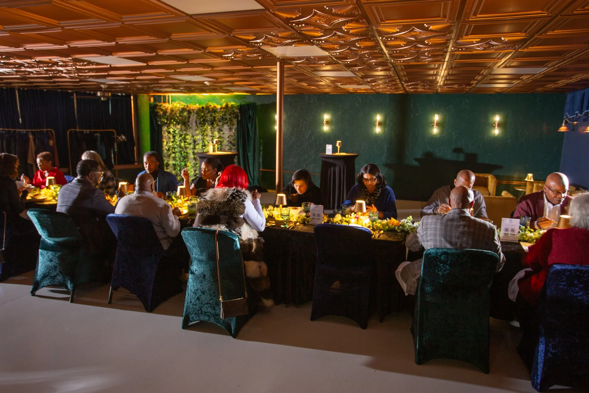 People dining at a long, elegant banquet table with dark tablecloth, decorated with flowers and small lamps, in a dimly lit, stylish event space with green walls and golden ceiling panels.