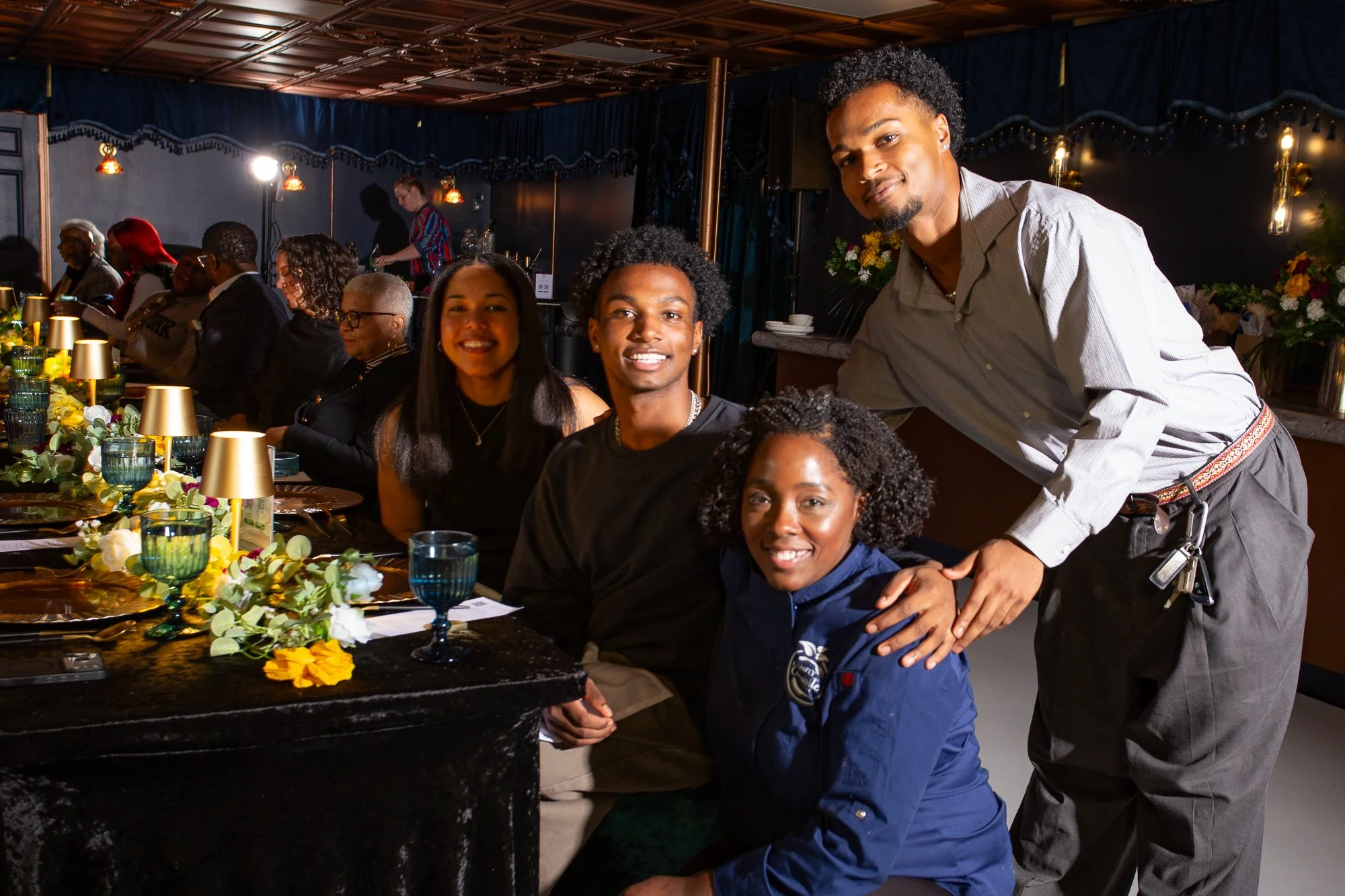People dining at a restaurant with a group photo of four individuals in the foreground and other diners seated along a decorated table.