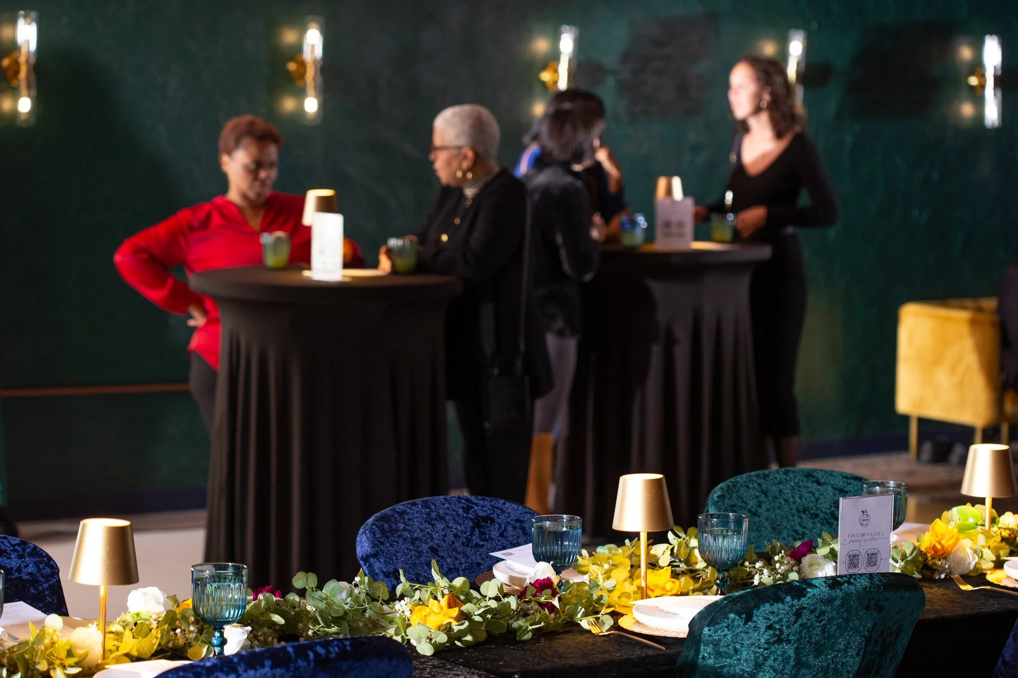 Decorated banquet table with blue and green chairs, floral centerpieces, glassware, and table lamps, in a dimly lit event space with standing high-top tables and guests socializing in the background.