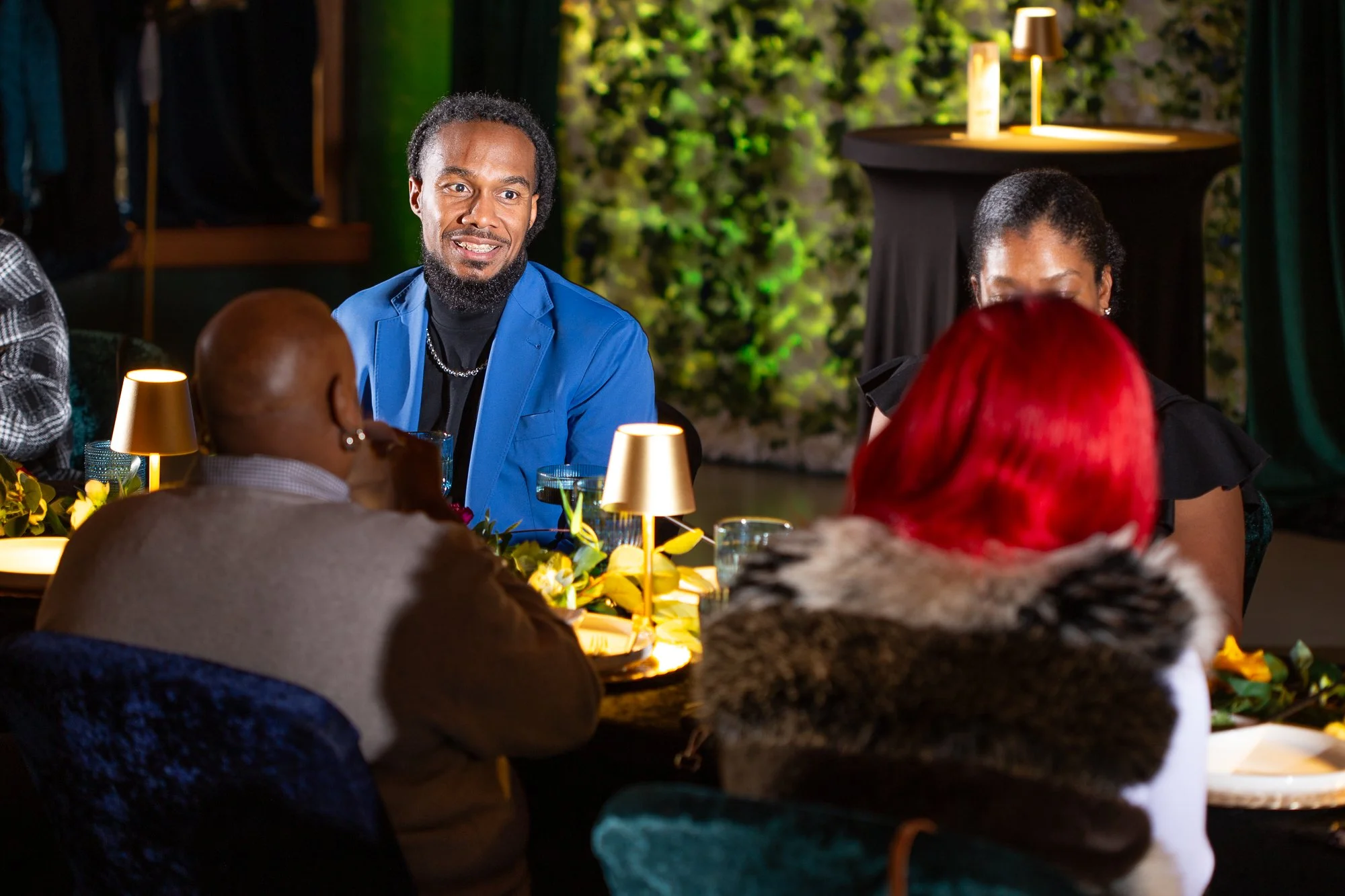 A group of people sitting around a dinner table in a dimly lit room, with a man in a blue jacket smiling and engaging with others, and a woman with red hair facing away. The table has candles and floral decorations.