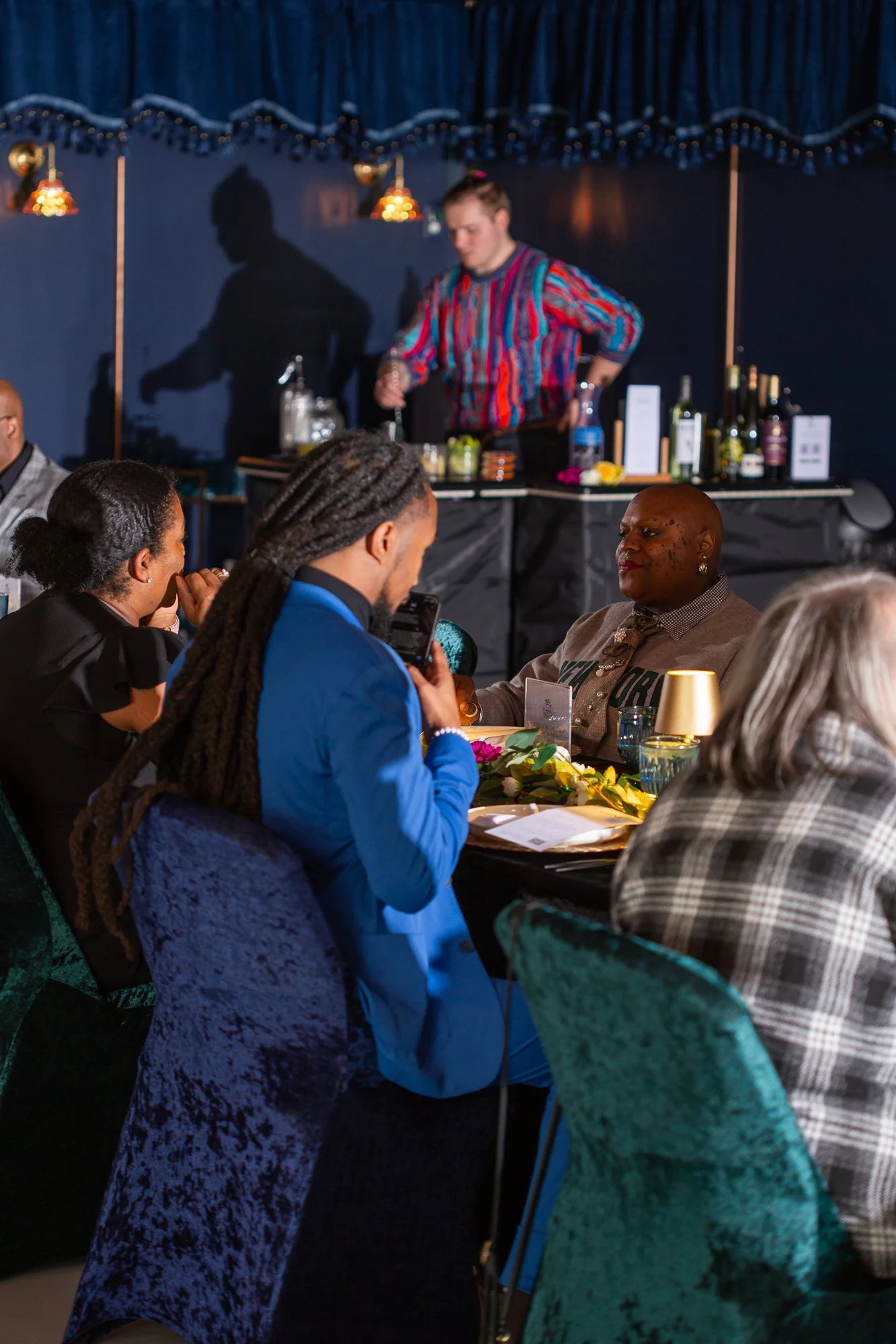 People seated at a dining event, with a bartender preparing drinks in the background, and a woman in a colorful striped shirt behind the bar.