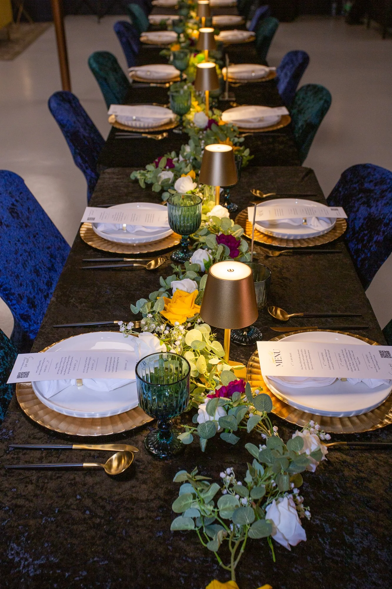 A banquet table set for a formal event with dark tablecloth, gold chargers, white plates, gold utensils, green glasses, small table lamps, and a floral center runner with yellow, white, and purple flowers and greenery.