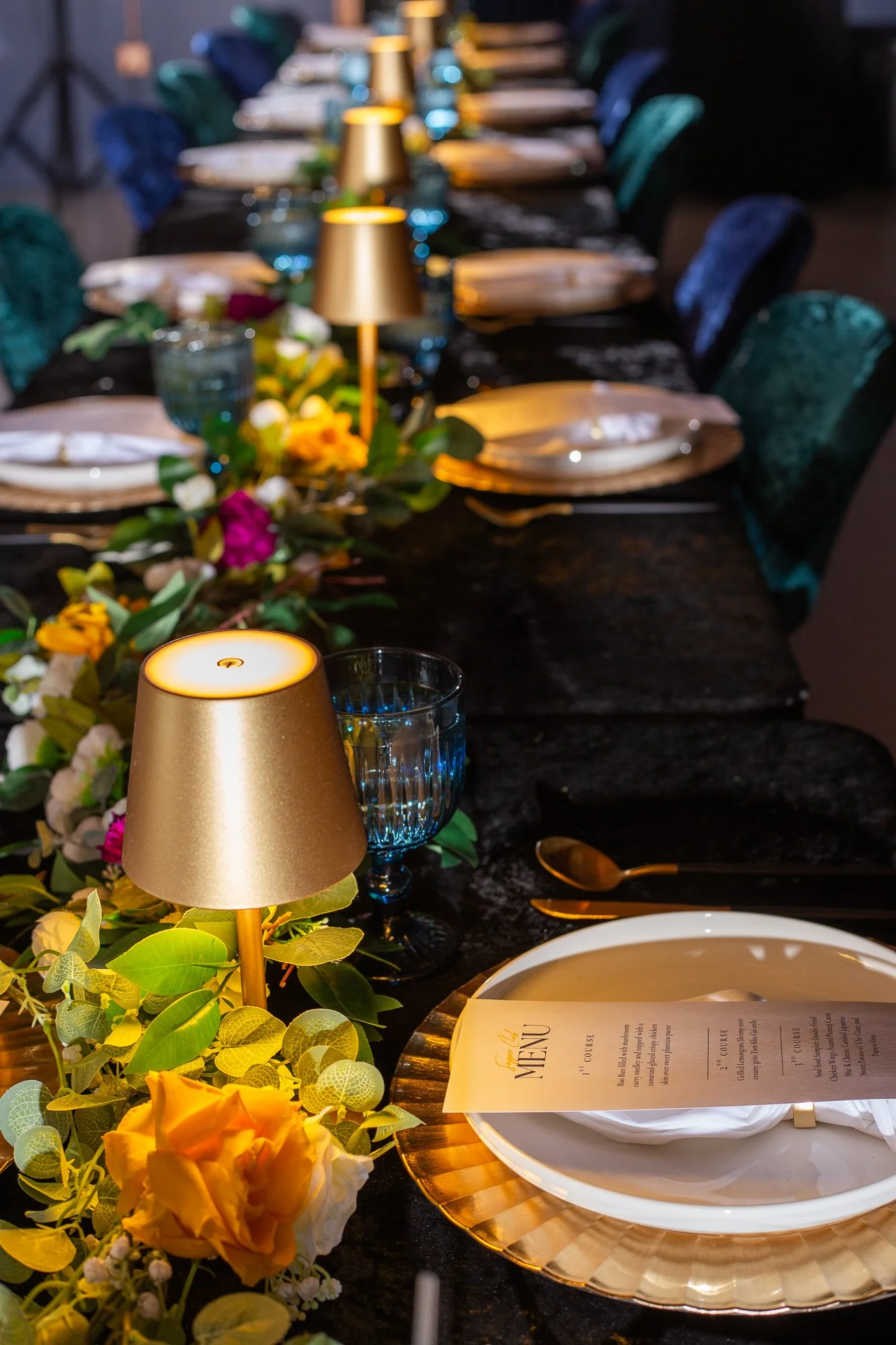 Elegant dining table with gold placemats, white plates, gold utensils, a menu, lantern-style candle holders, and colorful floral centerpieces, set for a formal event.