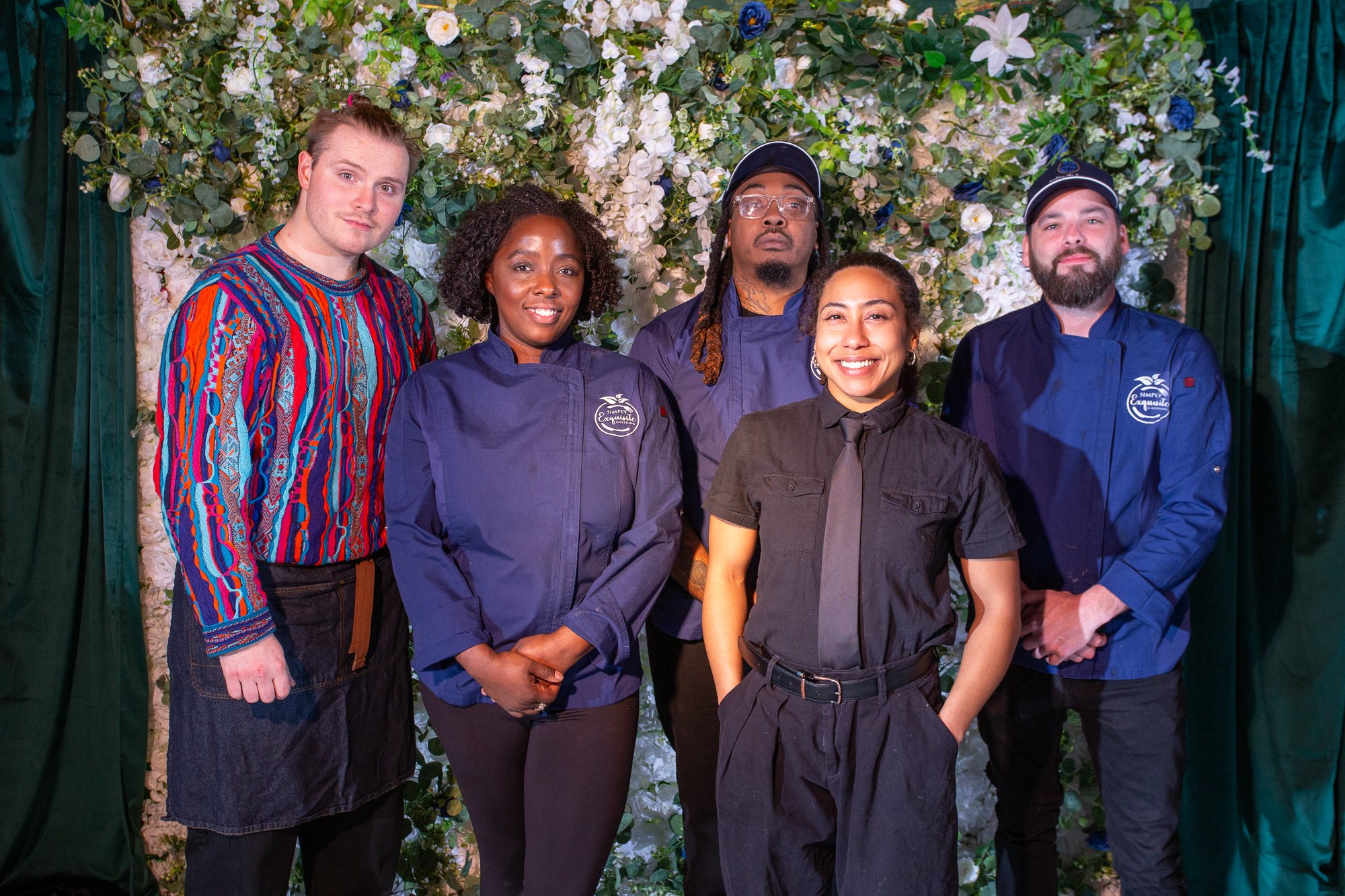 Group of five people standing in front of a floral backdrop, smiling at the camera, dressed in professional chef and server uniforms.