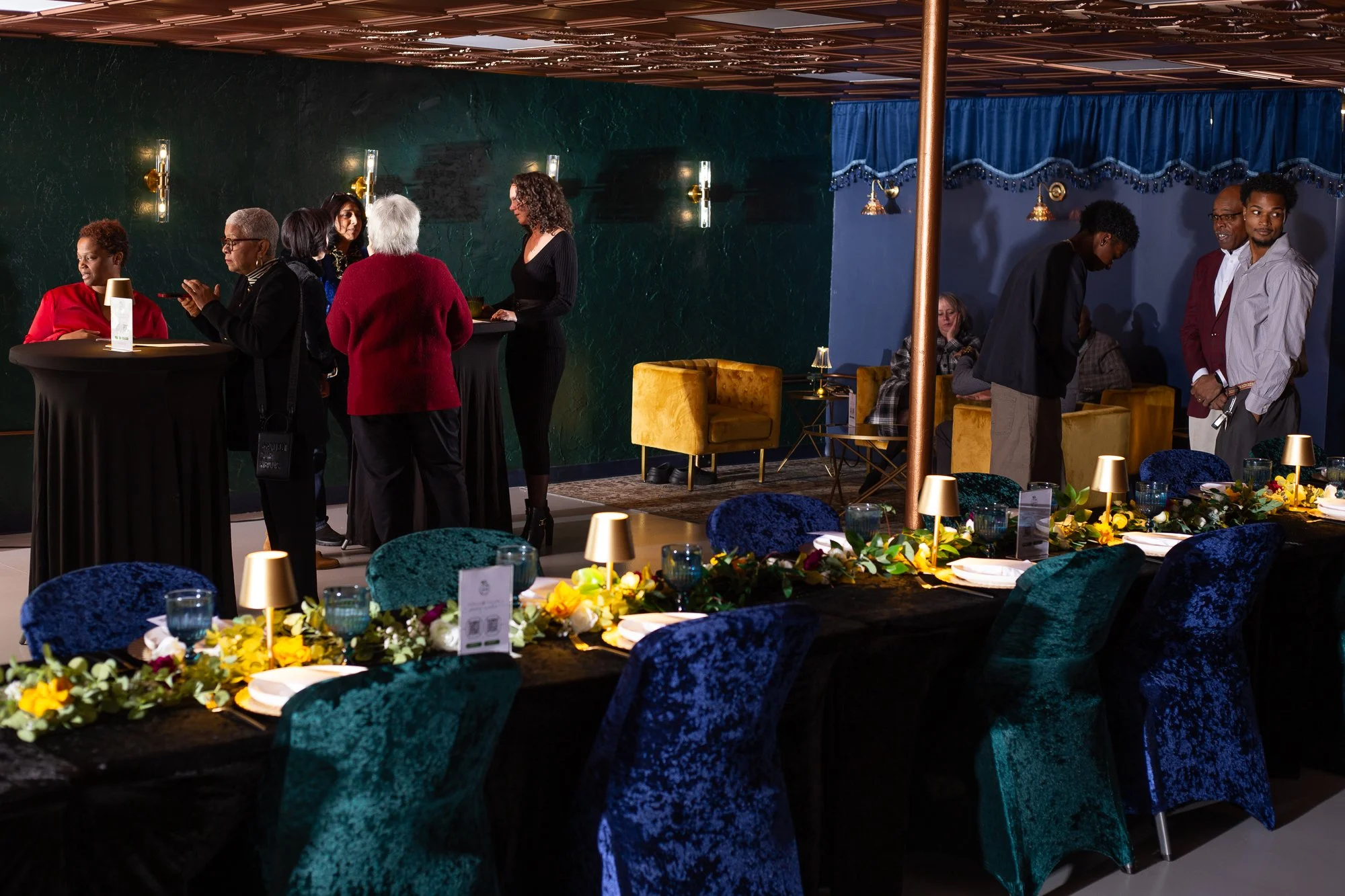 People socializing and sitting at tables in a dimly lit event space decorated with flowers and lamps.