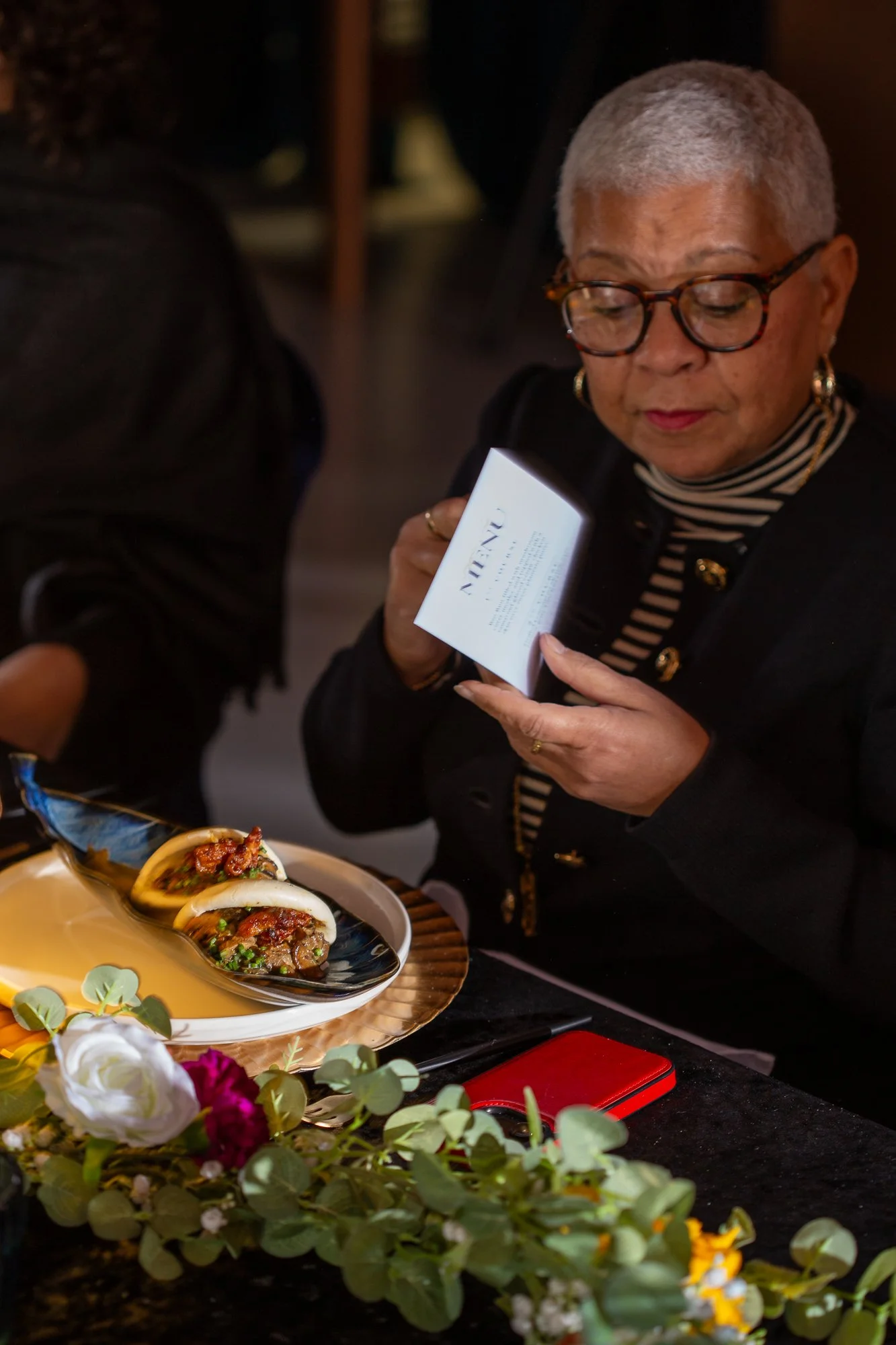 An elderly woman with short gray hair, glasses, and gold jewelry is looking at a menu while seated at a table with food. The table has a black tablecloth, and there are floral decorations and food items, including two small tacos on a black spoon and