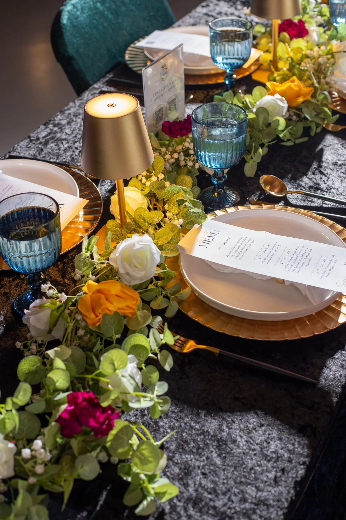A decorated dining table set for a meal with floral arrangements, blue water glasses, a menu, a gold-colored lamp, and gold cutlery, on a dark textured tablecloth.