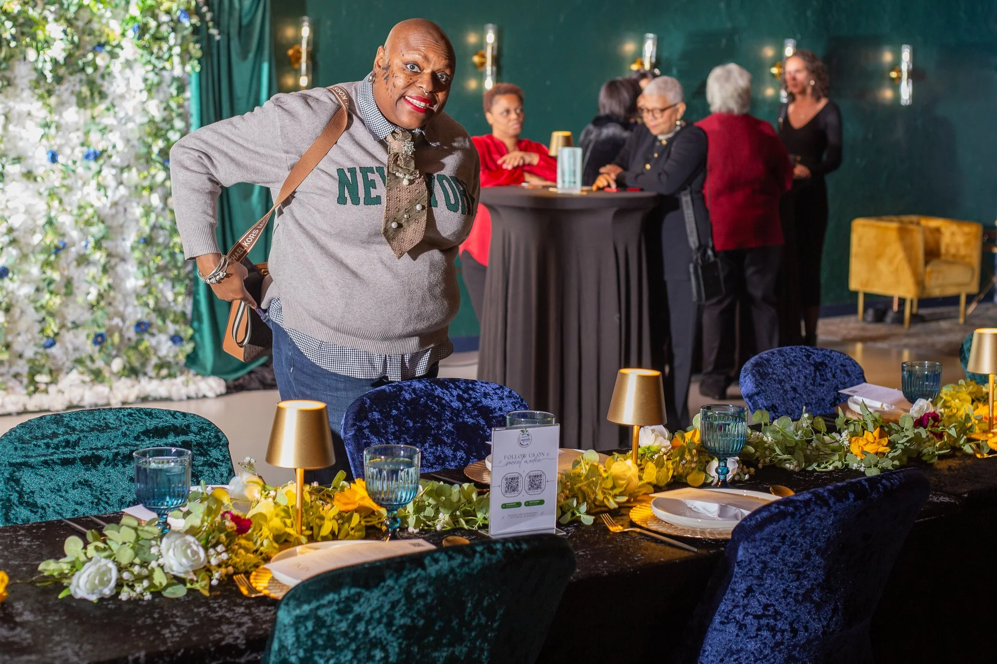A woman stands in front of a decorated table at an event, with a group of people conversing in the background.