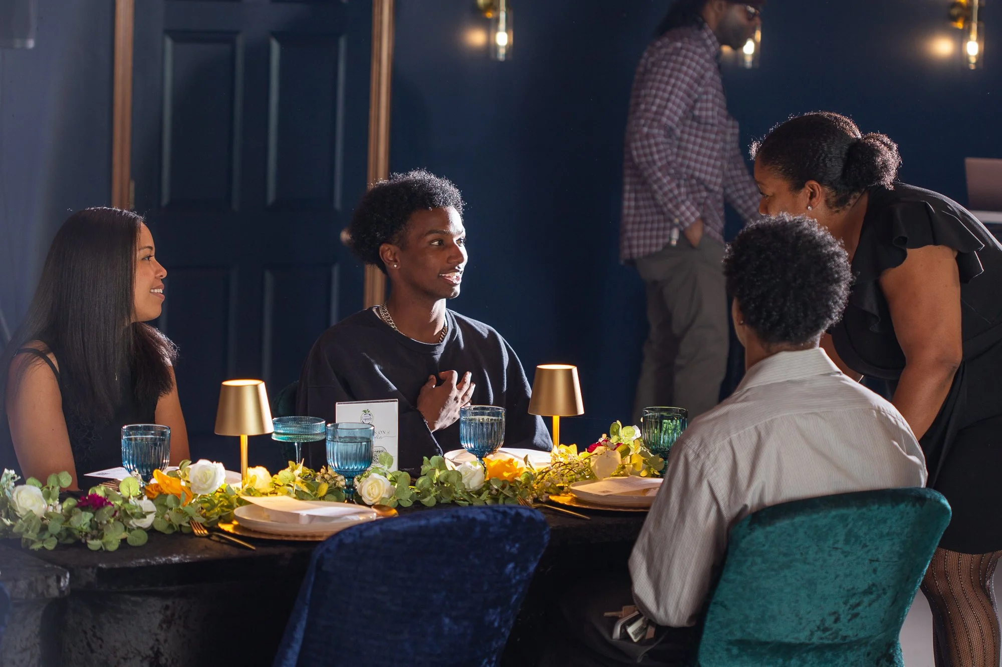 A group of four people having a conversation at a decorated dining table in a dimly lit room, with one woman standing and leaning towards the group.