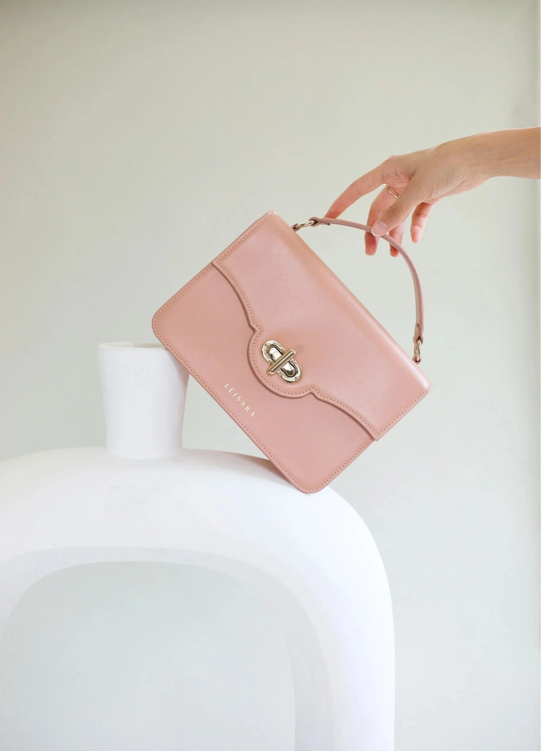 A pink handbag with a silver clasp being held by a woman's hand, positioned above a white, modern, curved furniture piece.