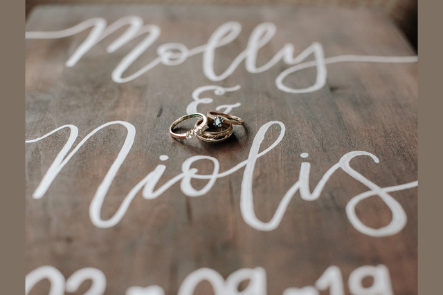 Close-up of wedding rings on a custom hand-lettered wooden sign with white cursive text that reads 'Molly & Niolis'. Sign is a keepsake or gift for the couple.